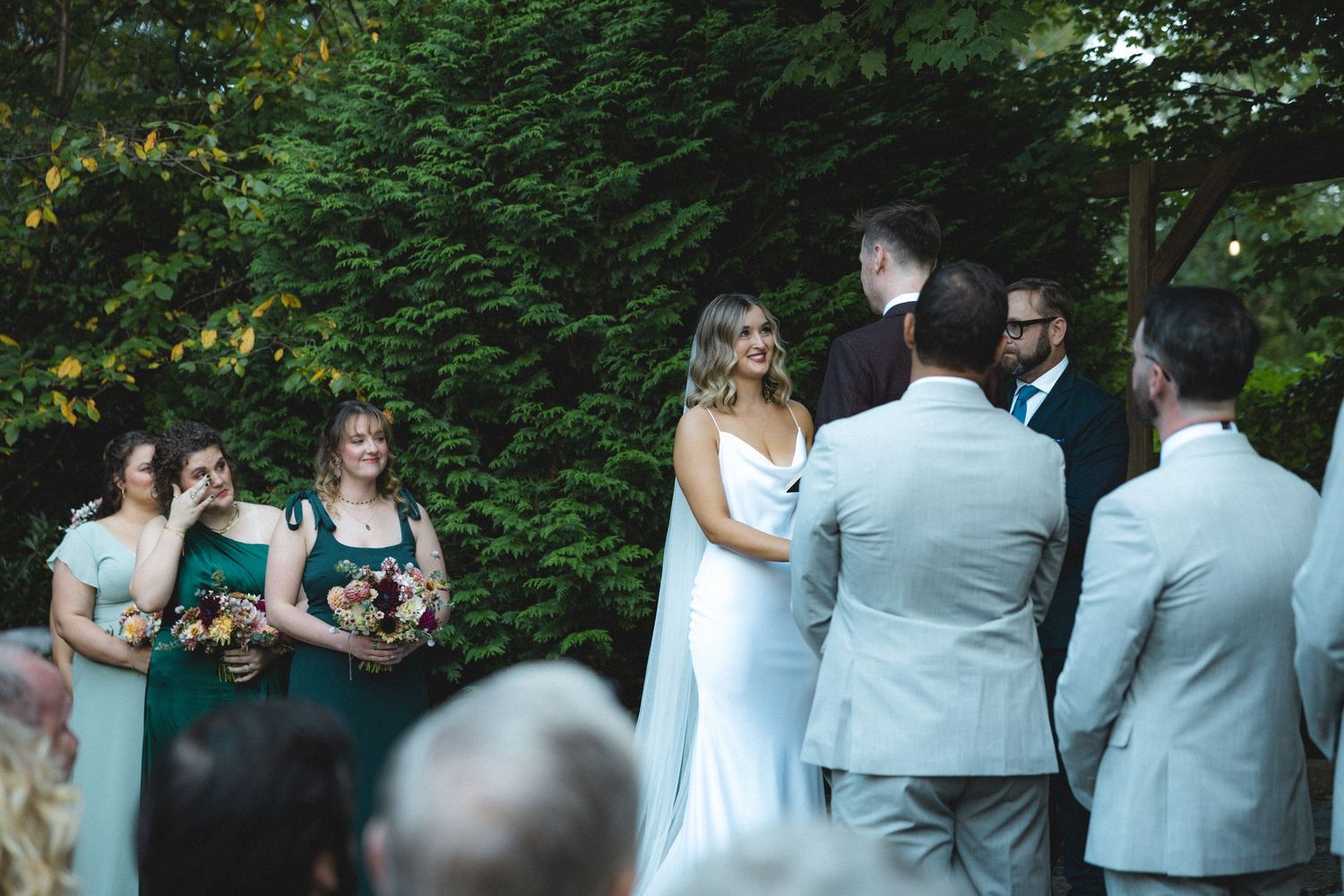 Outdoor wedding ceremony with bride in white gown, bridesmaids in green dresses, and guests seated among lush greenery.