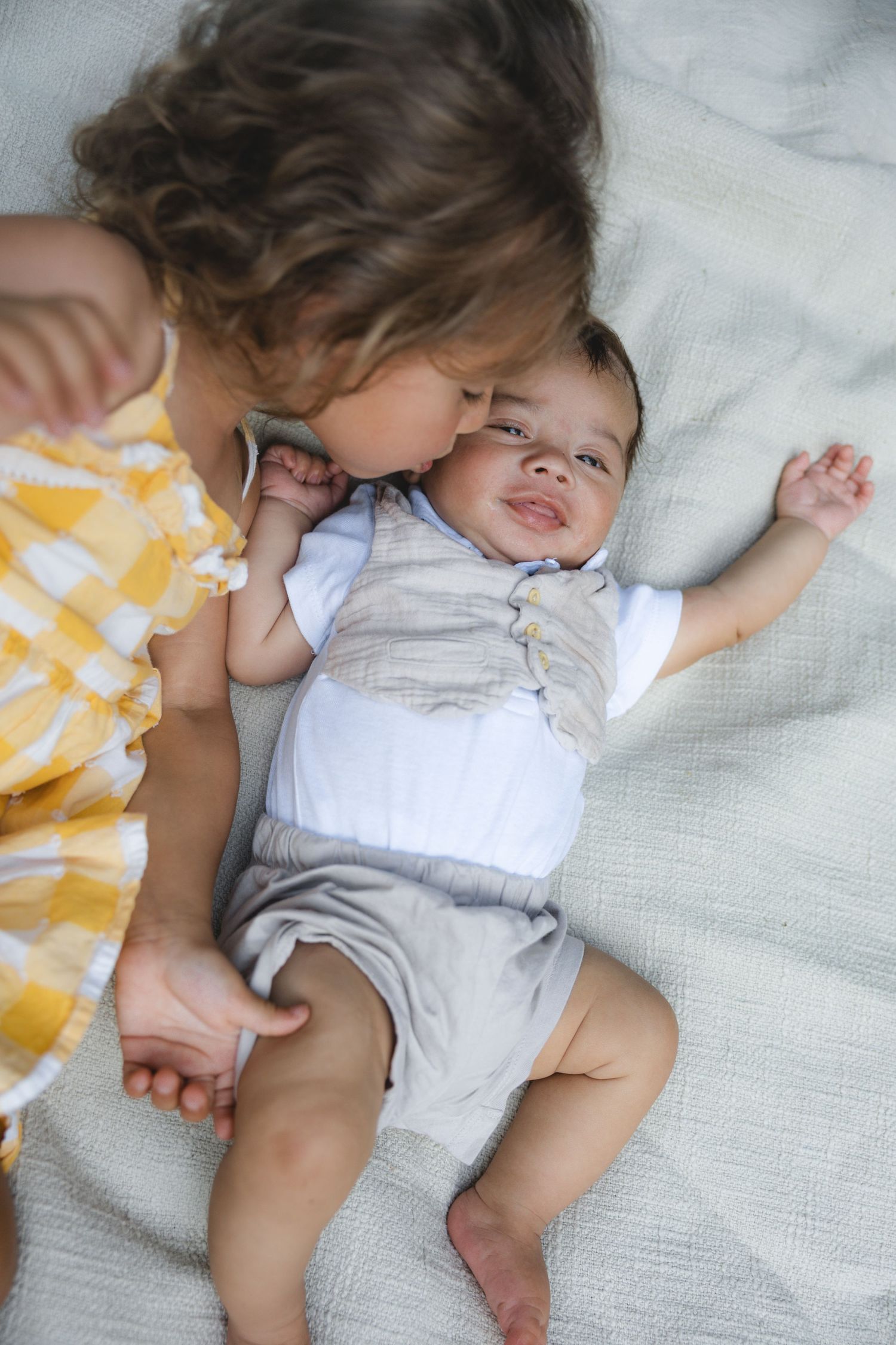 Young girl in yellow dress kissing smiling baby sibling lying on white blanket.