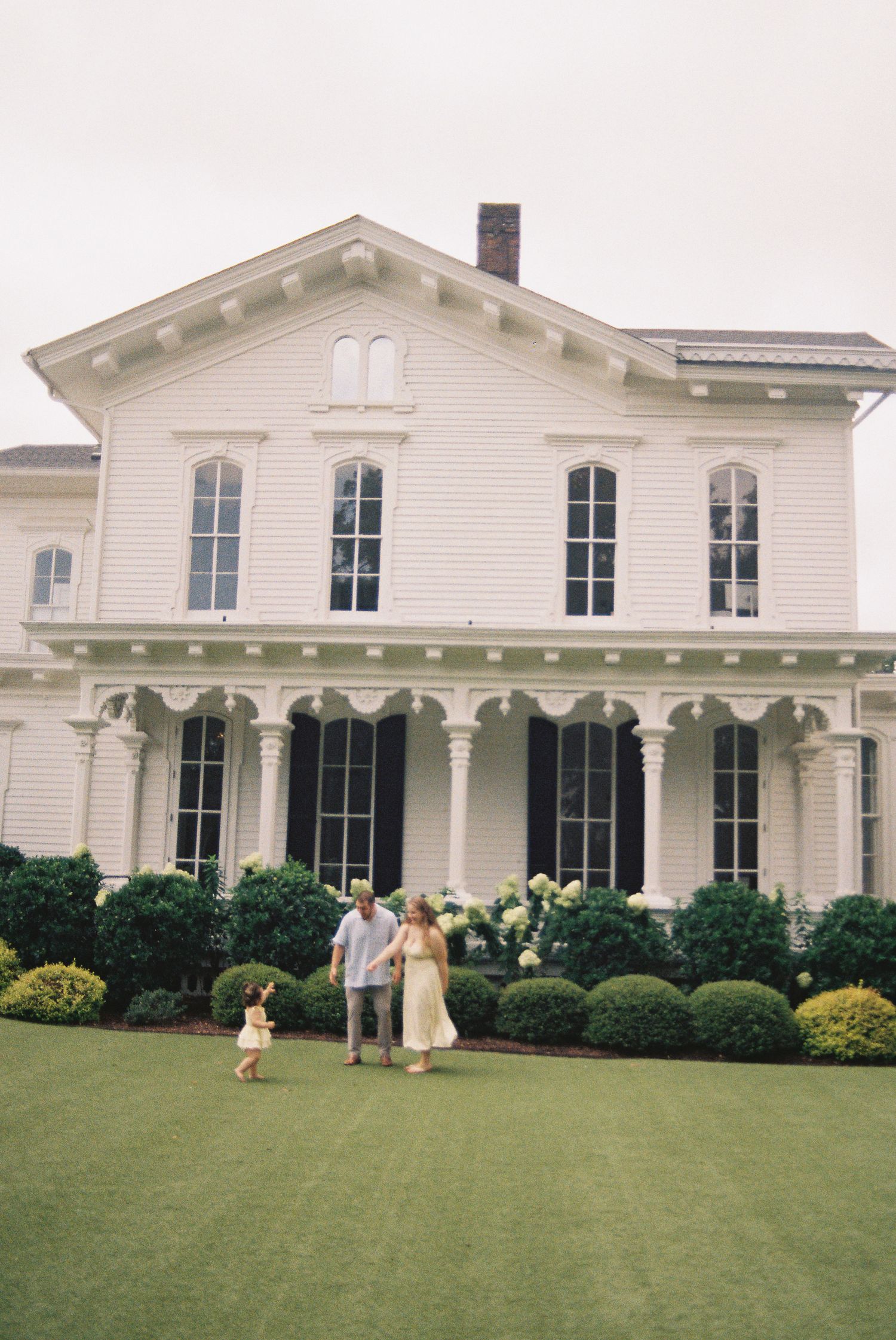 Family of three walking on green lawn before grand white Southern colonial home with black shutters.