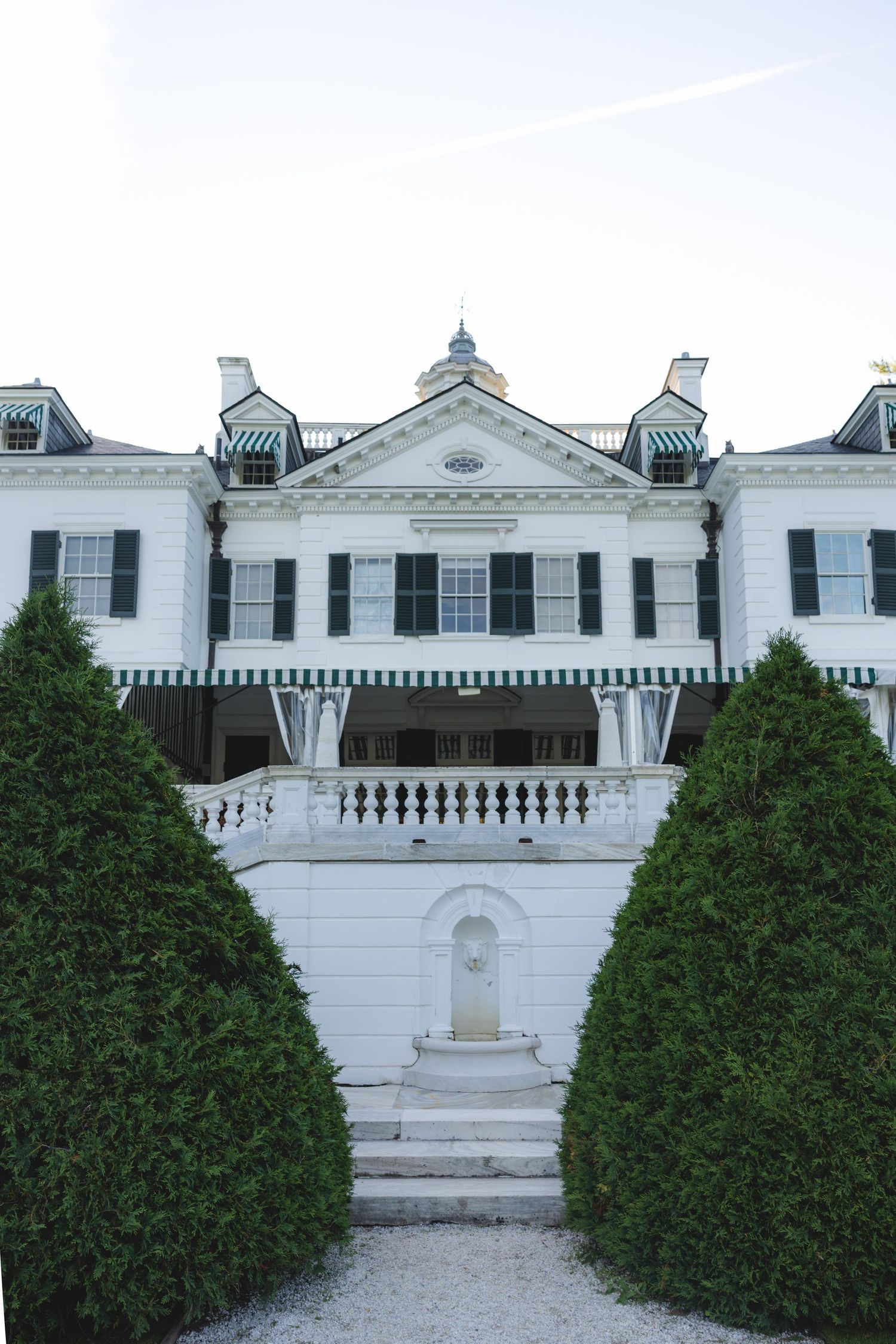 White colonial mansion with green striped awnings, black shutters, manicured hedges, and a grand staircase leading to the entrance.