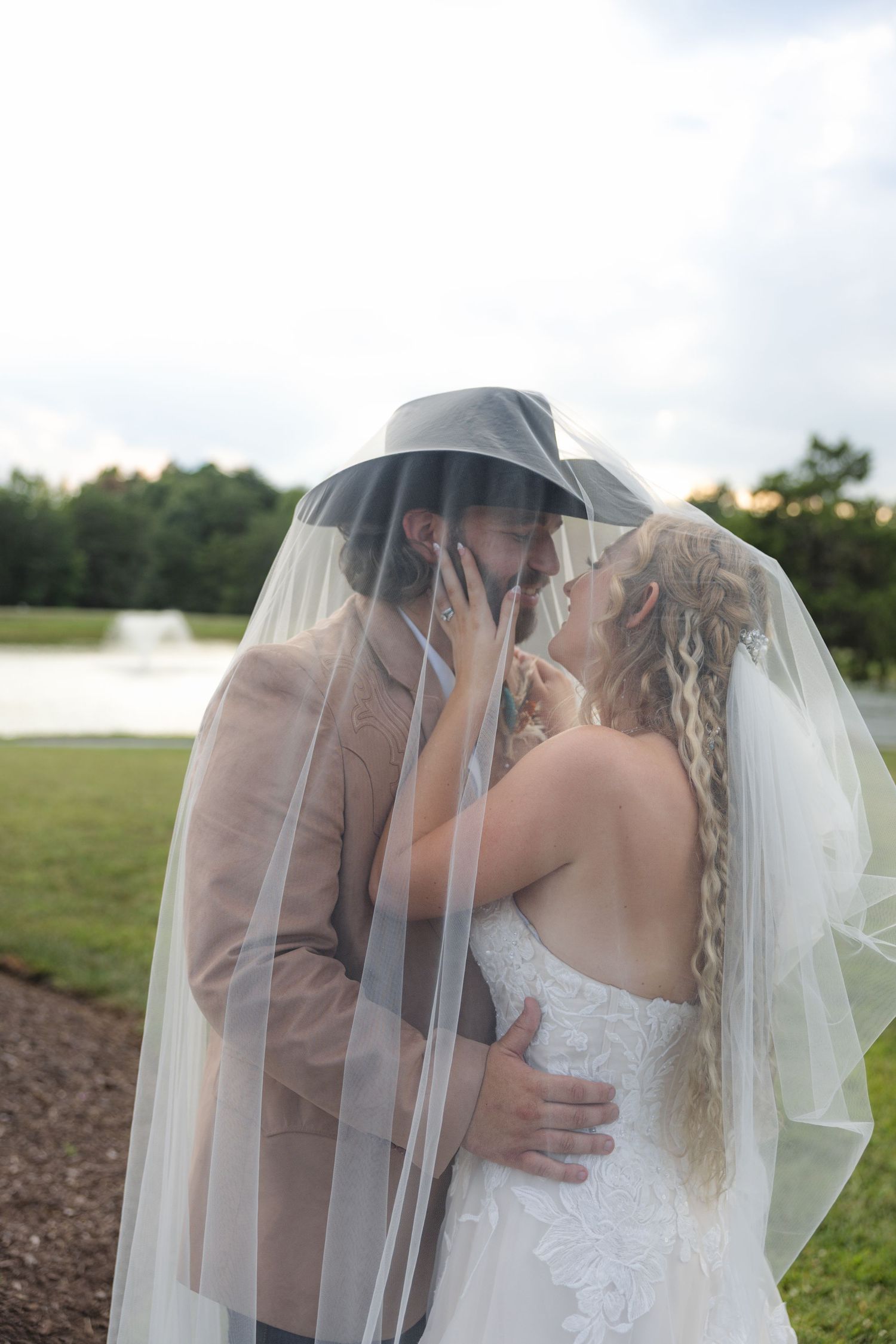 Bride and groom kissing under a wedding veil outdoors, groom wearing a cowboy hat and tan suit.