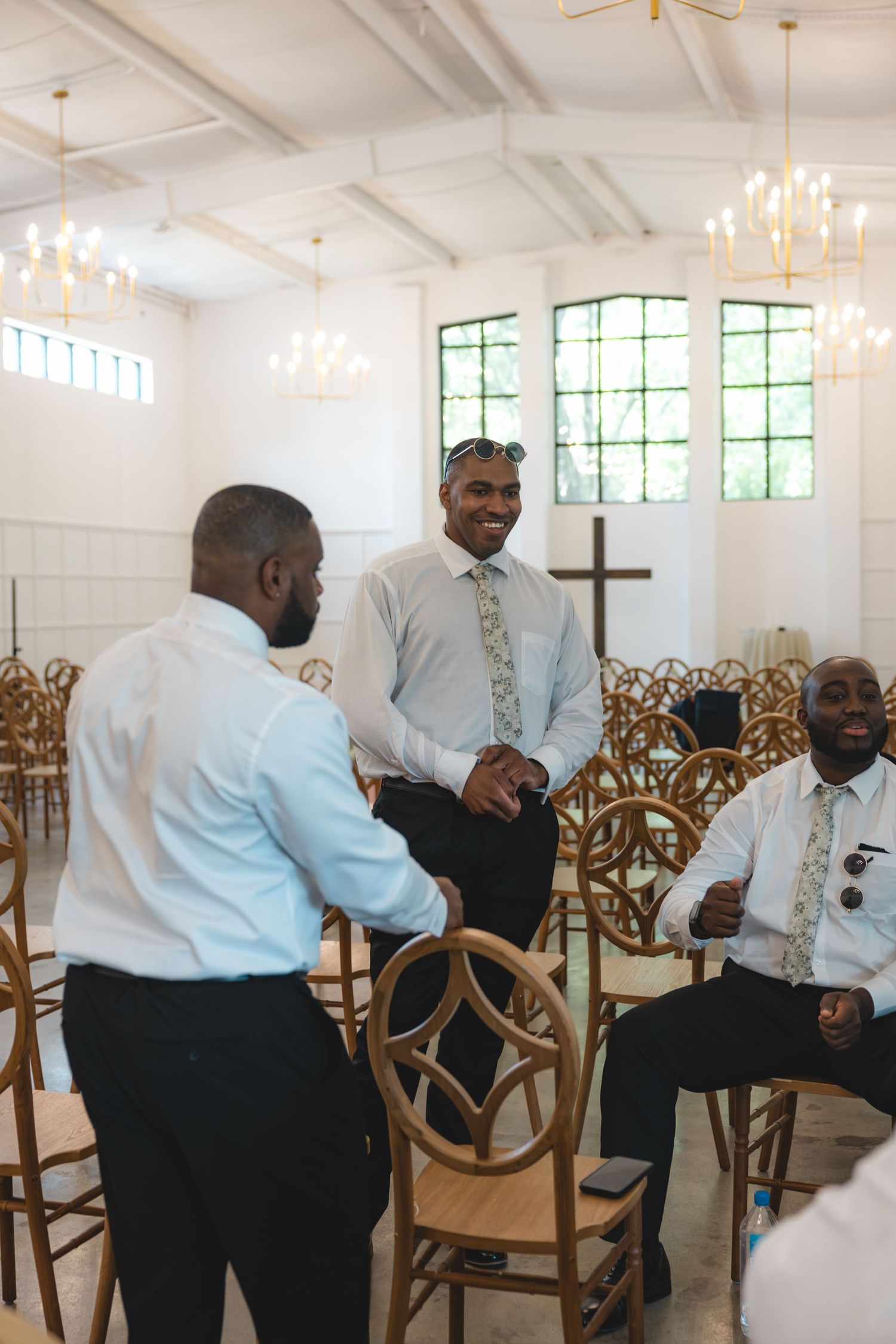 Men in dress shirts laughing and socializing inside a bright white wedding chapel with wooden chairs and a cross.