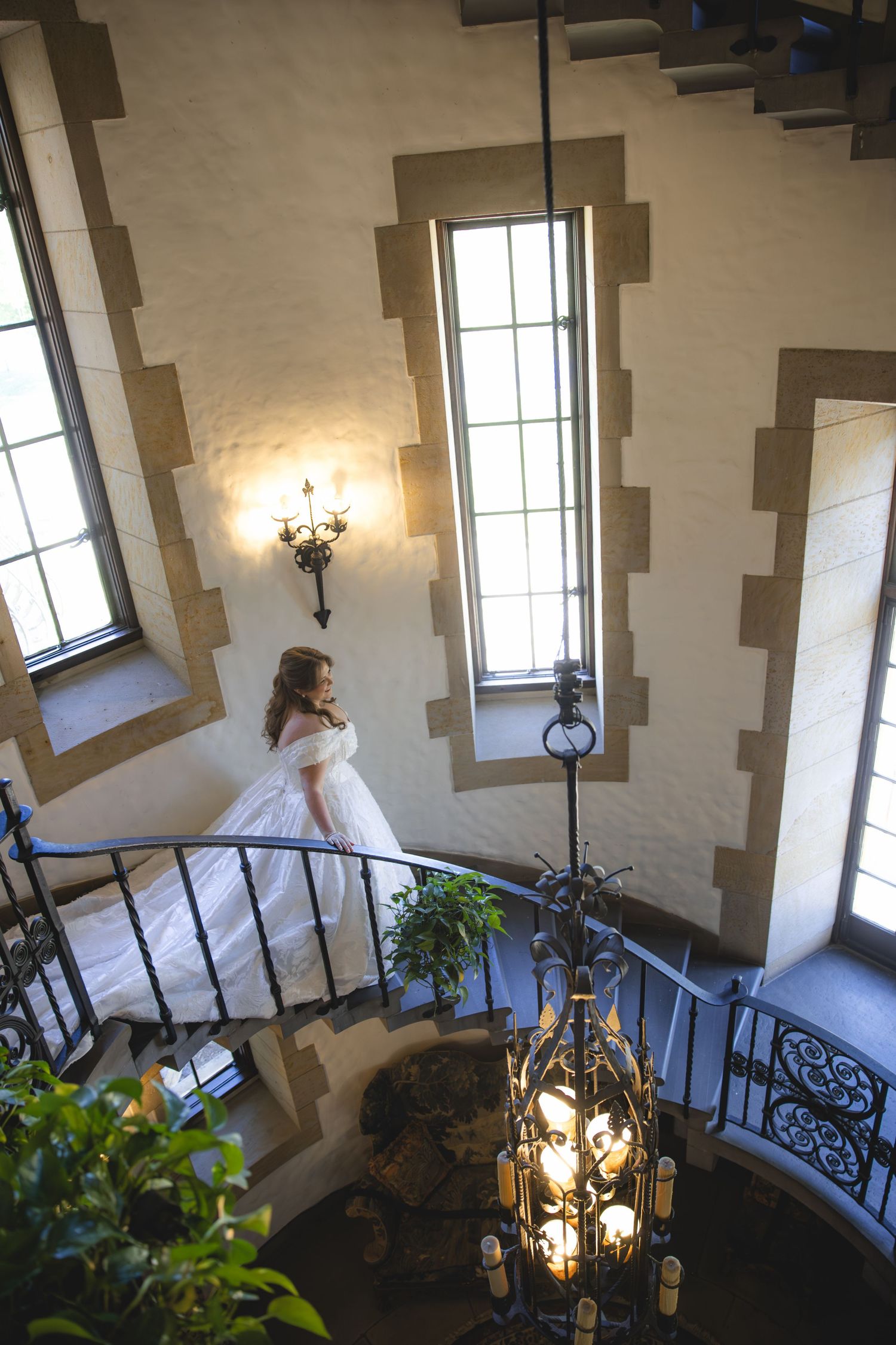 Bride in white gown on spiral staircase in castle-like venue with chandelier and tall windows.