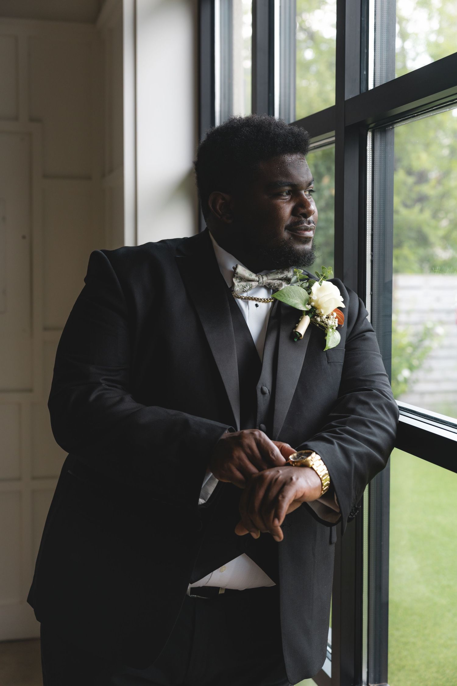 A well-dressed groom in a black tuxedo with bow tie and boutonniere gazes thoughtfully out a large window.