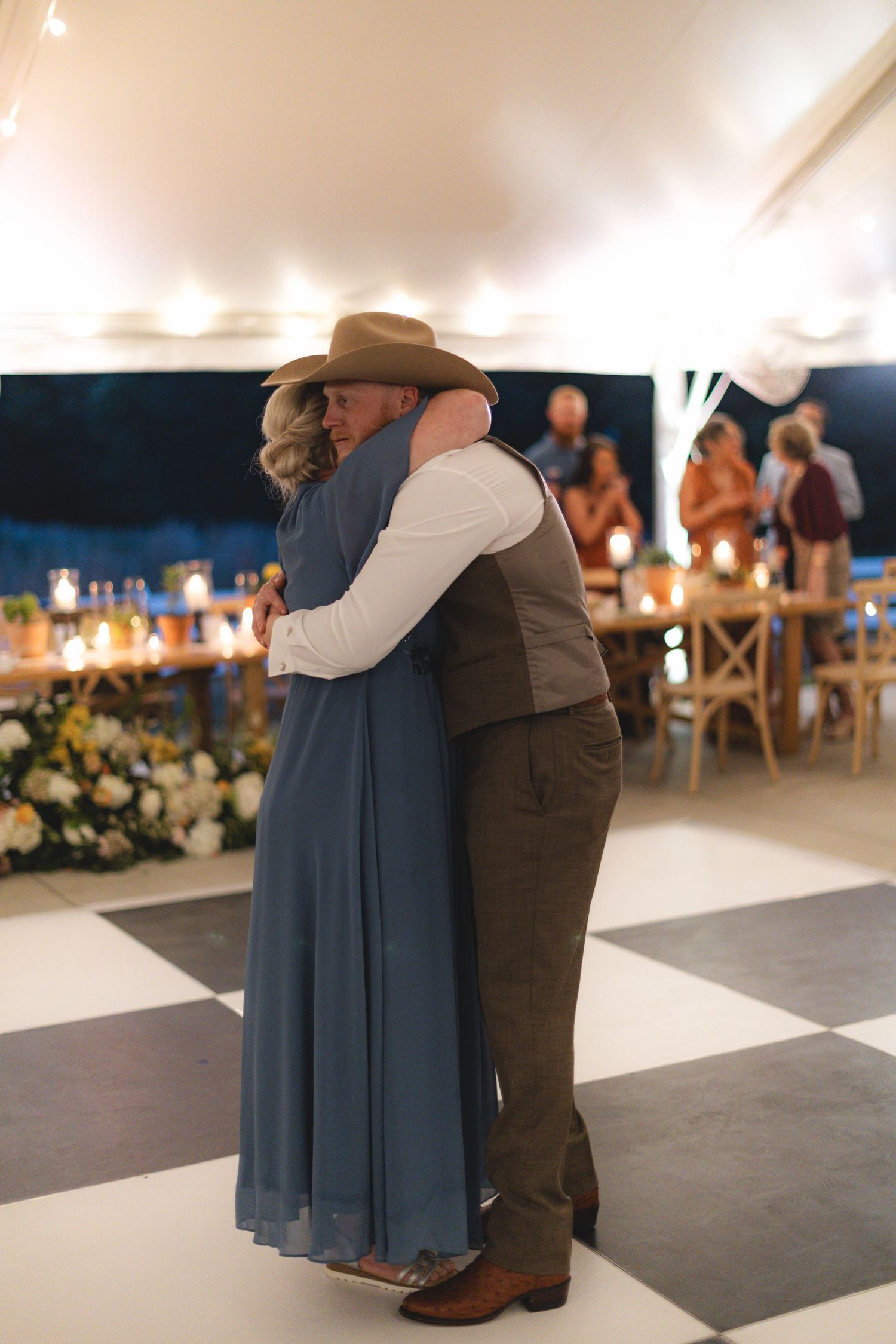 Groom in cowboy hat and brown vest shares tender mother-son dance at rustic wedding reception.