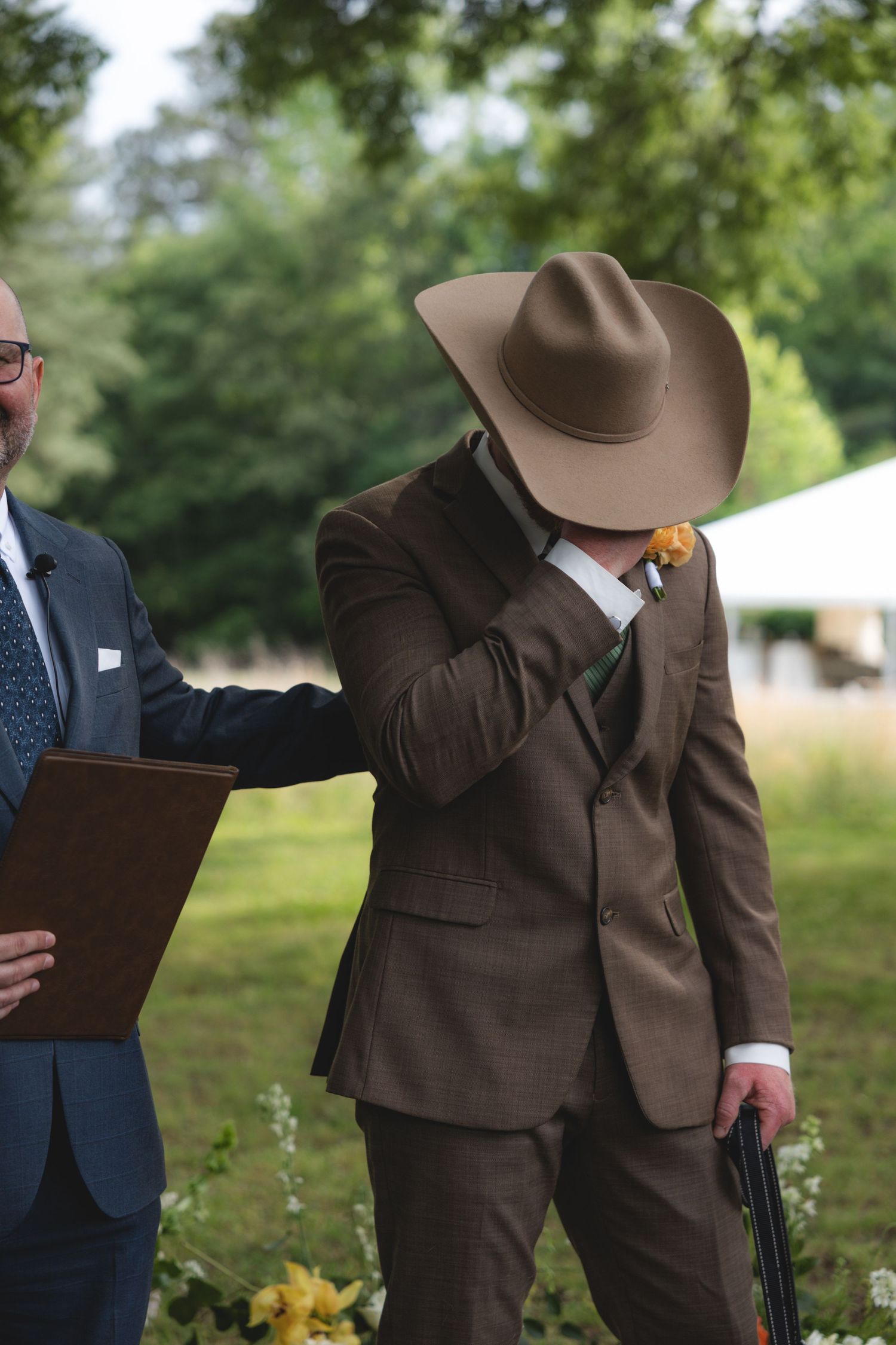 Emotional groom in brown suit and cowboy hat wiping tears during outdoor wedding ceremony.