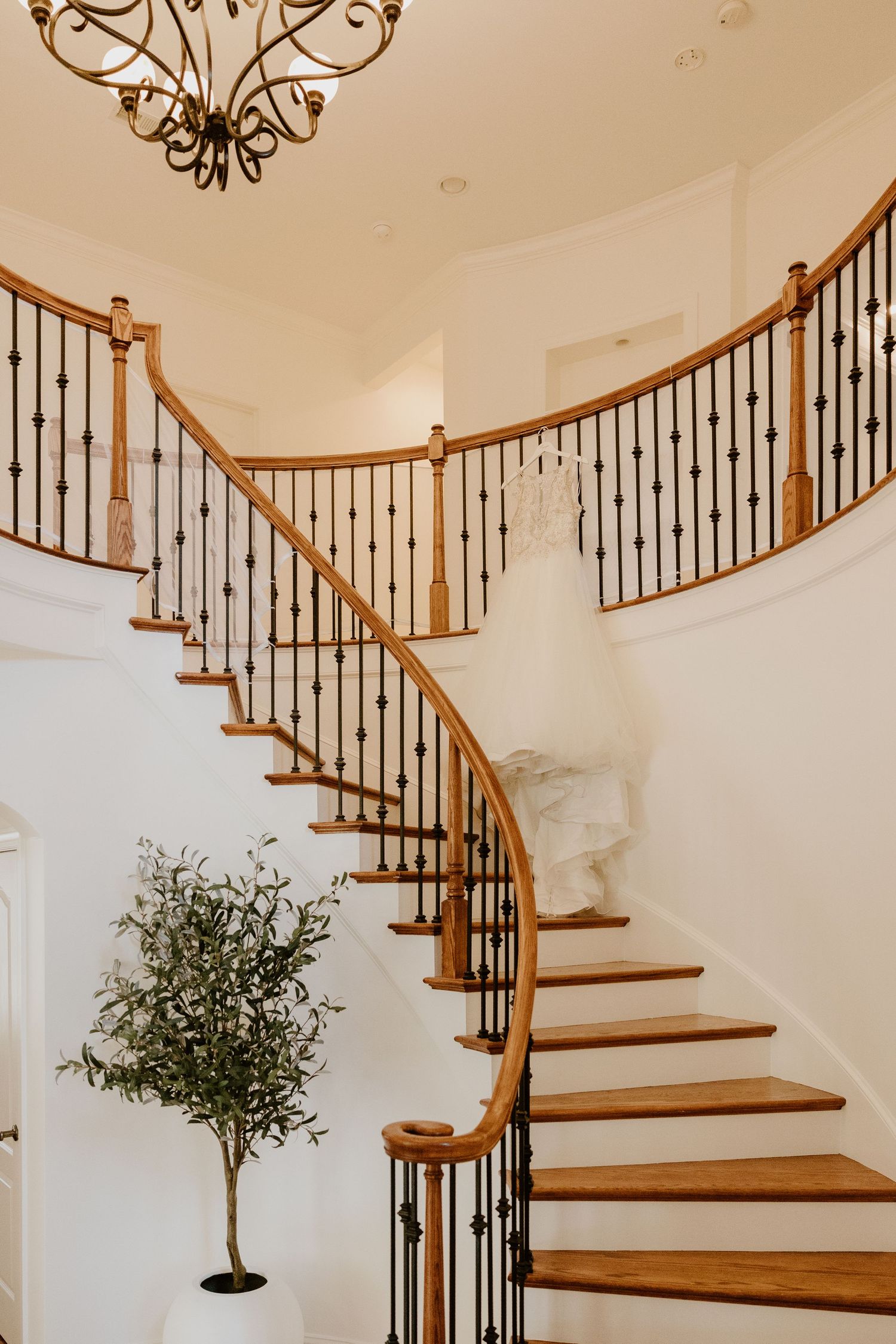Elegant white wedding gown draped over curved wooden staircase railing with wrought iron balusters and chandelier above.