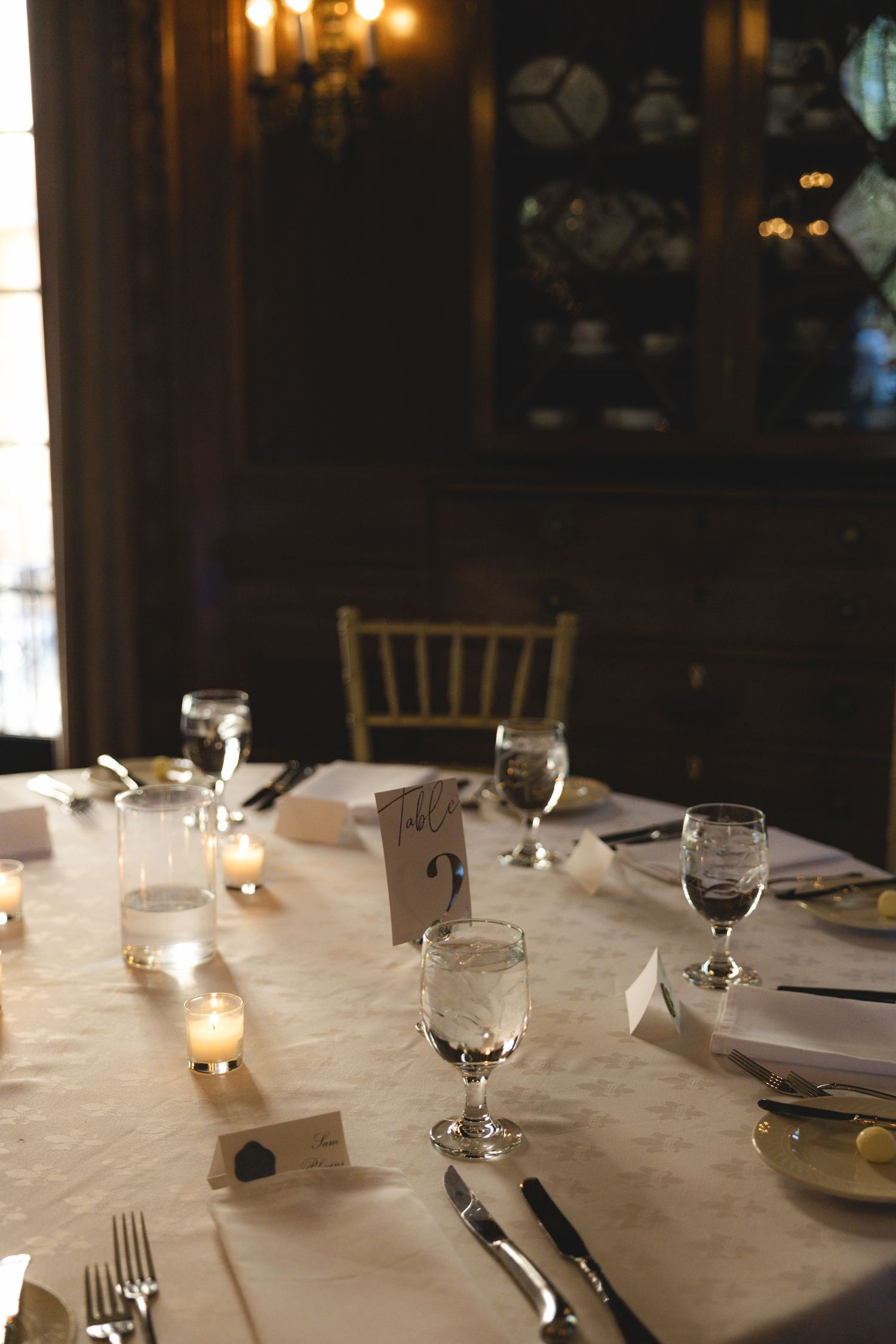 Elegant wedding reception table set with white linen, candles, crystal glassware, and a table number card in a dimly lit venue.