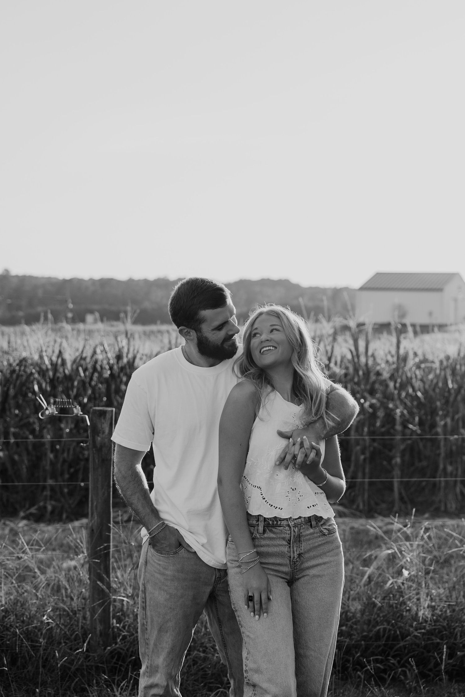 Black and white photo of a smiling couple embracing near a fence with a cornfield and barn in the background.
