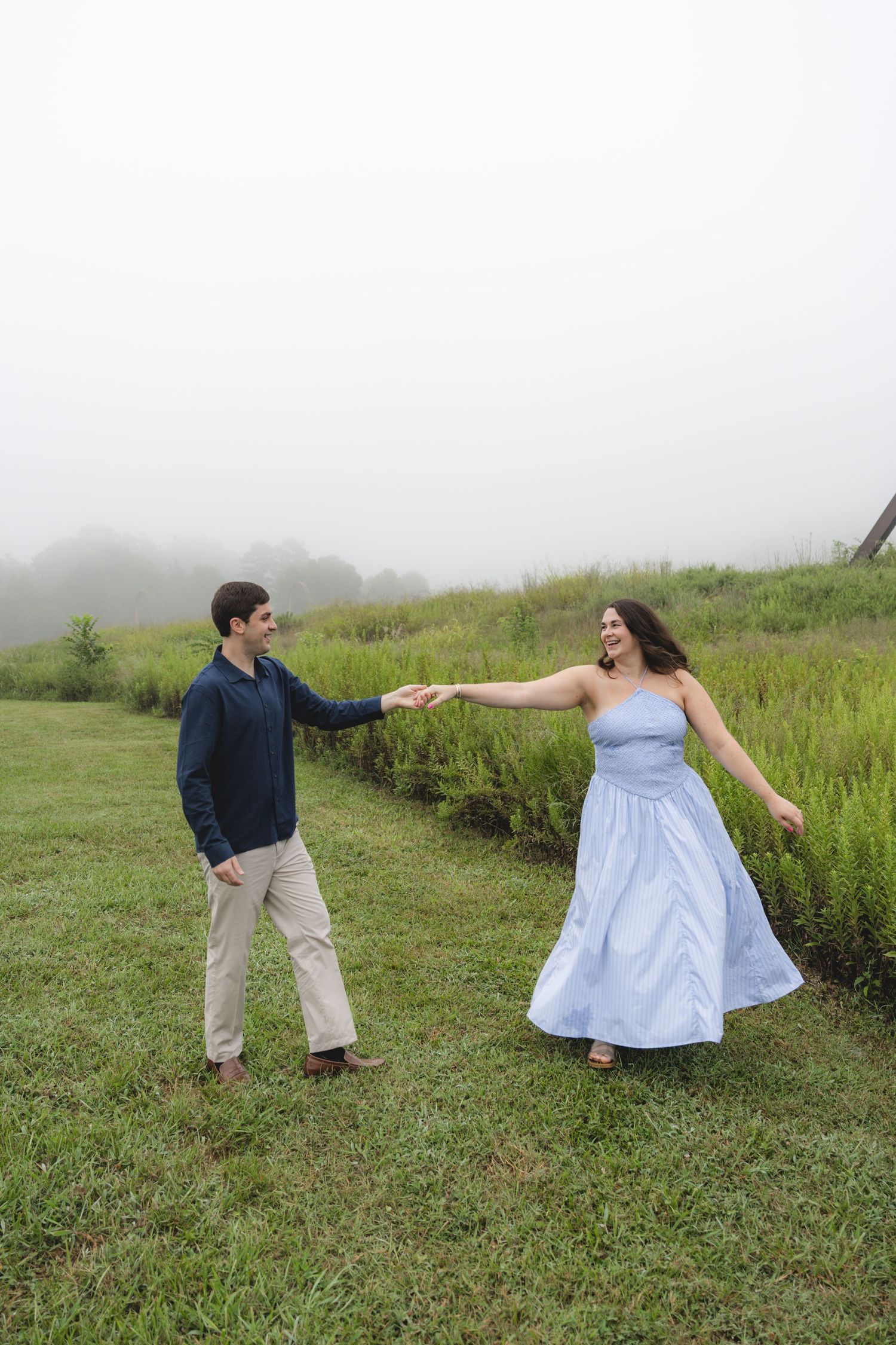 A couple dances joyfully in a misty green field, the woman wearing a flowing light blue dress holding hands with a man in navy.