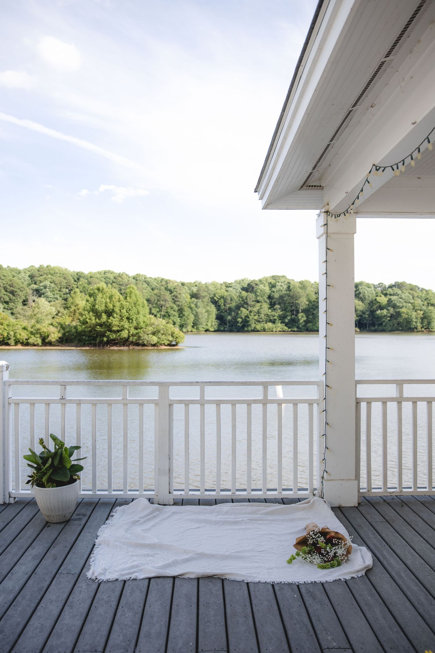 White deck overlooking a serene lake with lush green trees, featuring a white blanket, flowers, and a potted plant.