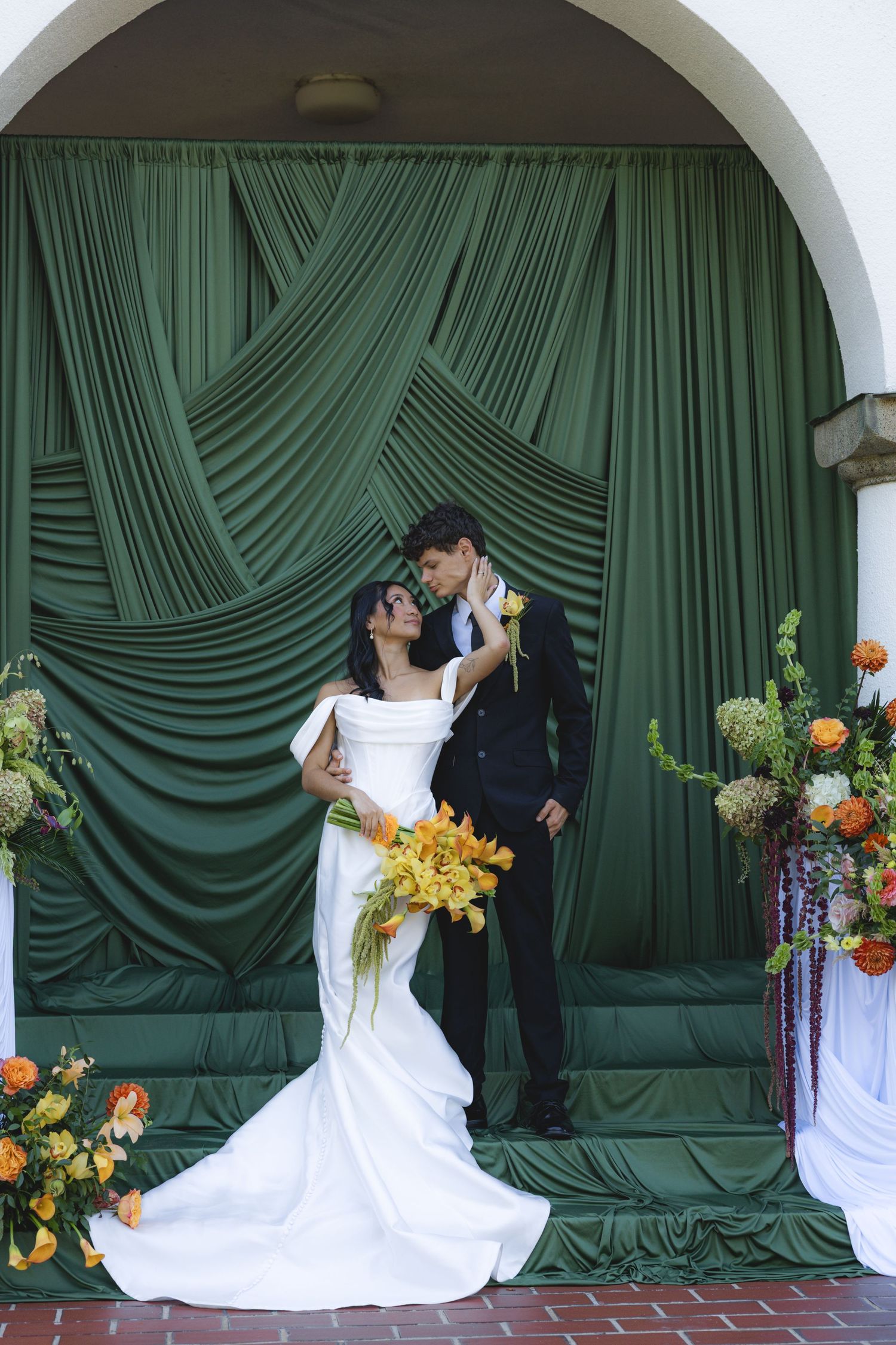 Bride in white mermaid gown and groom in black suit pose before green draped backdrop with yellow floral arrangements.