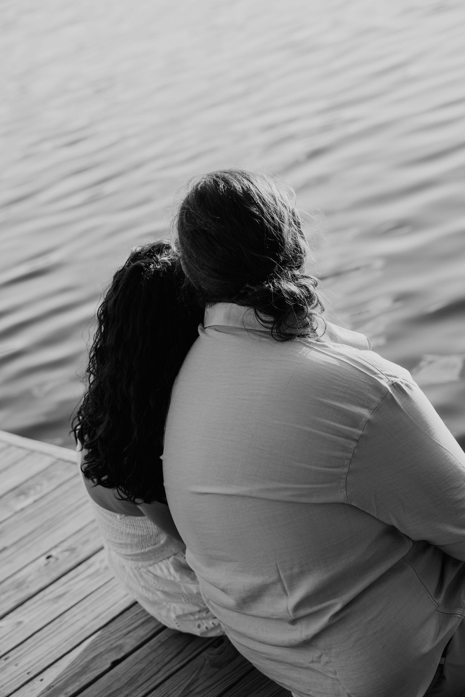 Black and white photo of a couple sitting on a dock, embracing while gazing at calm water.