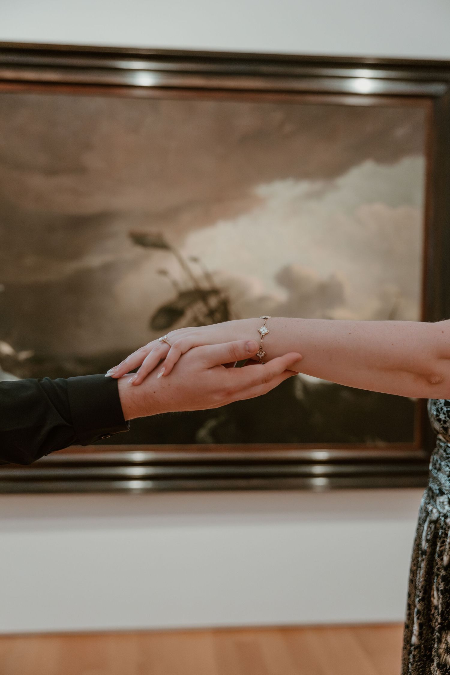 Two hands gently touching, showcasing a delicate diamond ring, with a classic framed painting in the background.