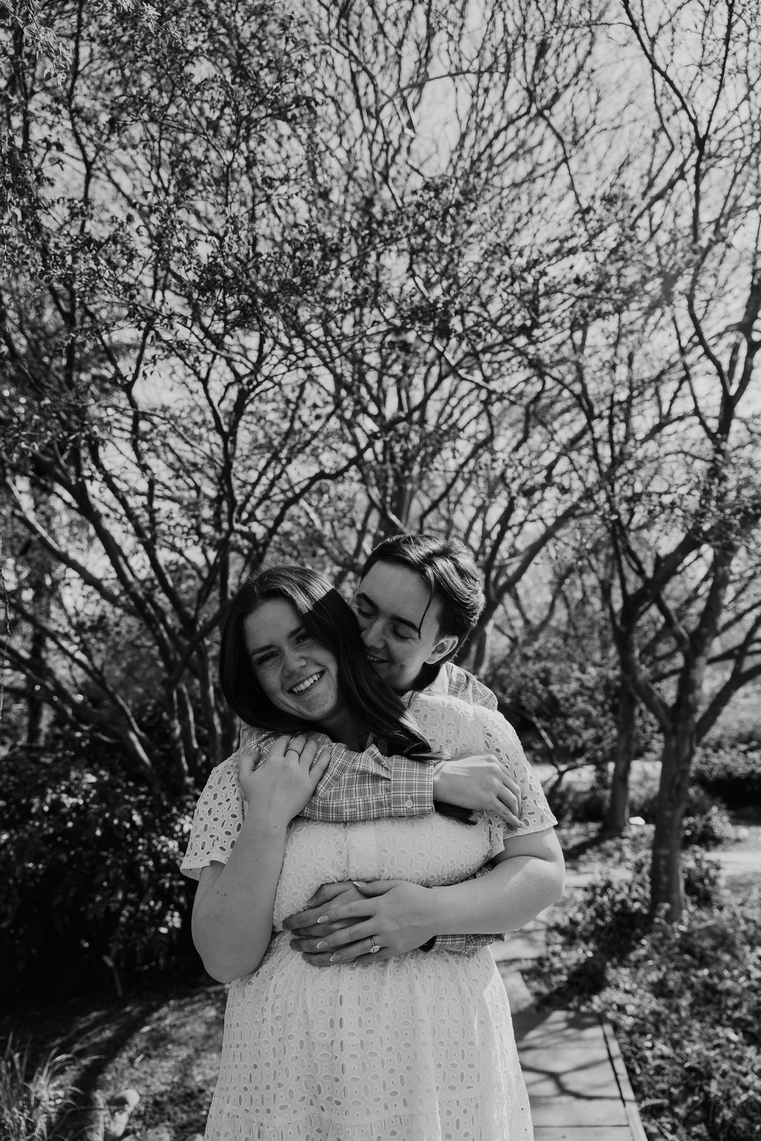 Black and white photo of a couple embracing outdoors, man hugging woman from behind among bare trees.