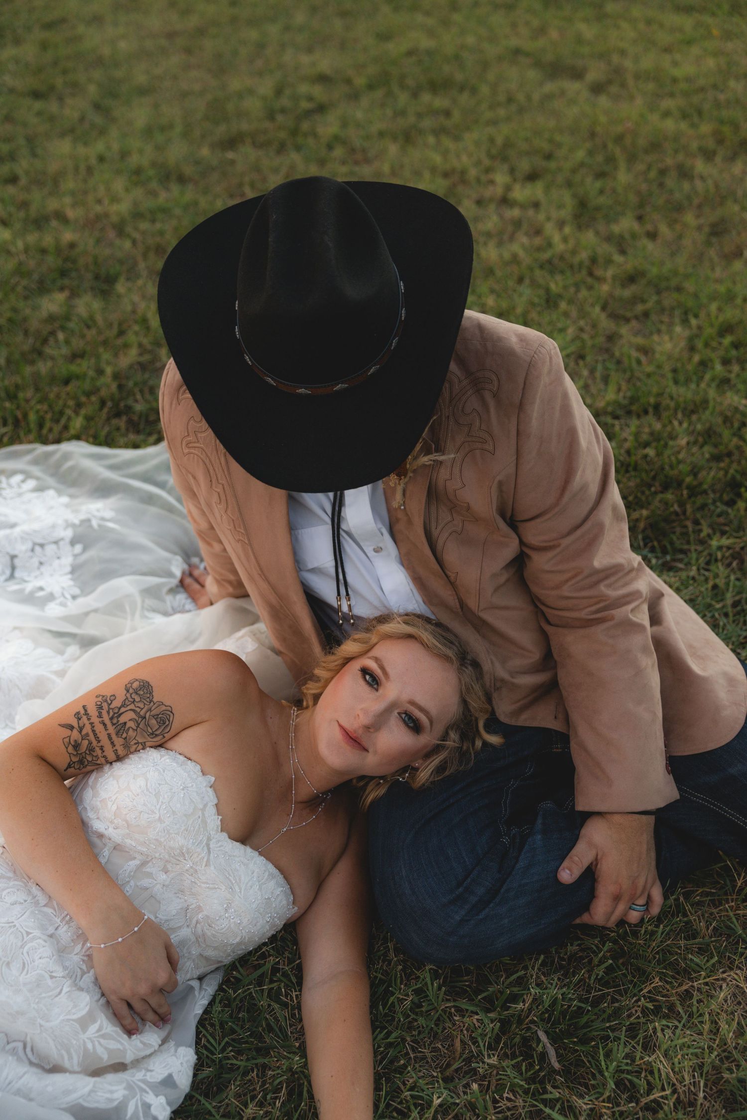 Bride in white gown lying on grass while groom in tan blazer and black cowboy hat leans over her outdoors.