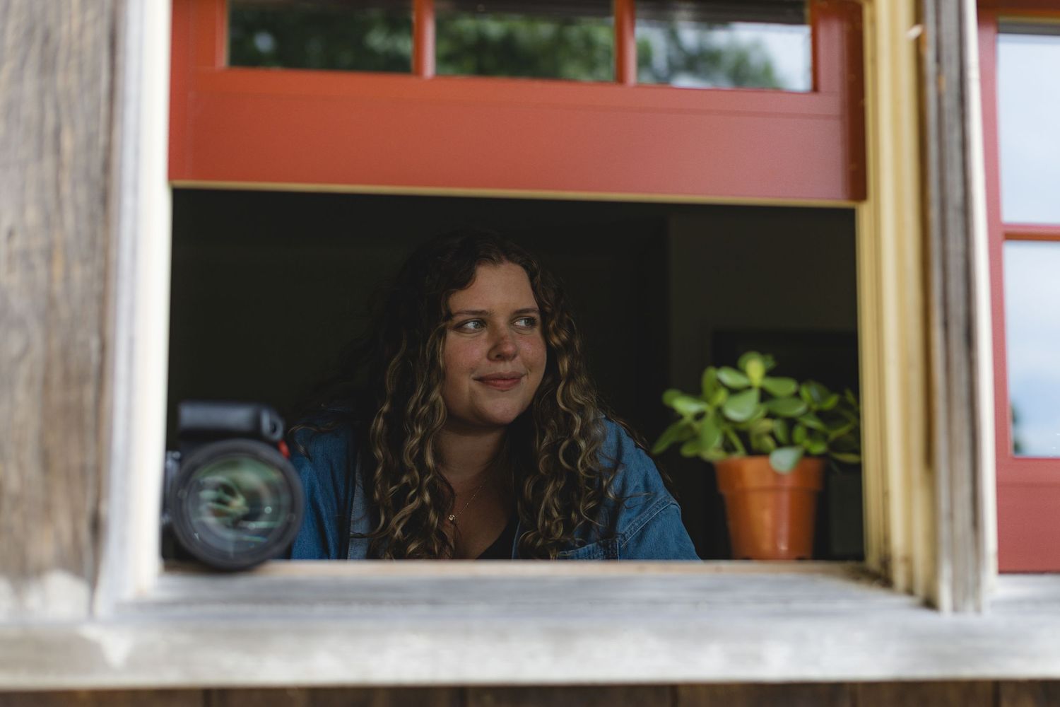Young woman with curly hair gazing upward through an open window with red trim and a potted plant nearby.