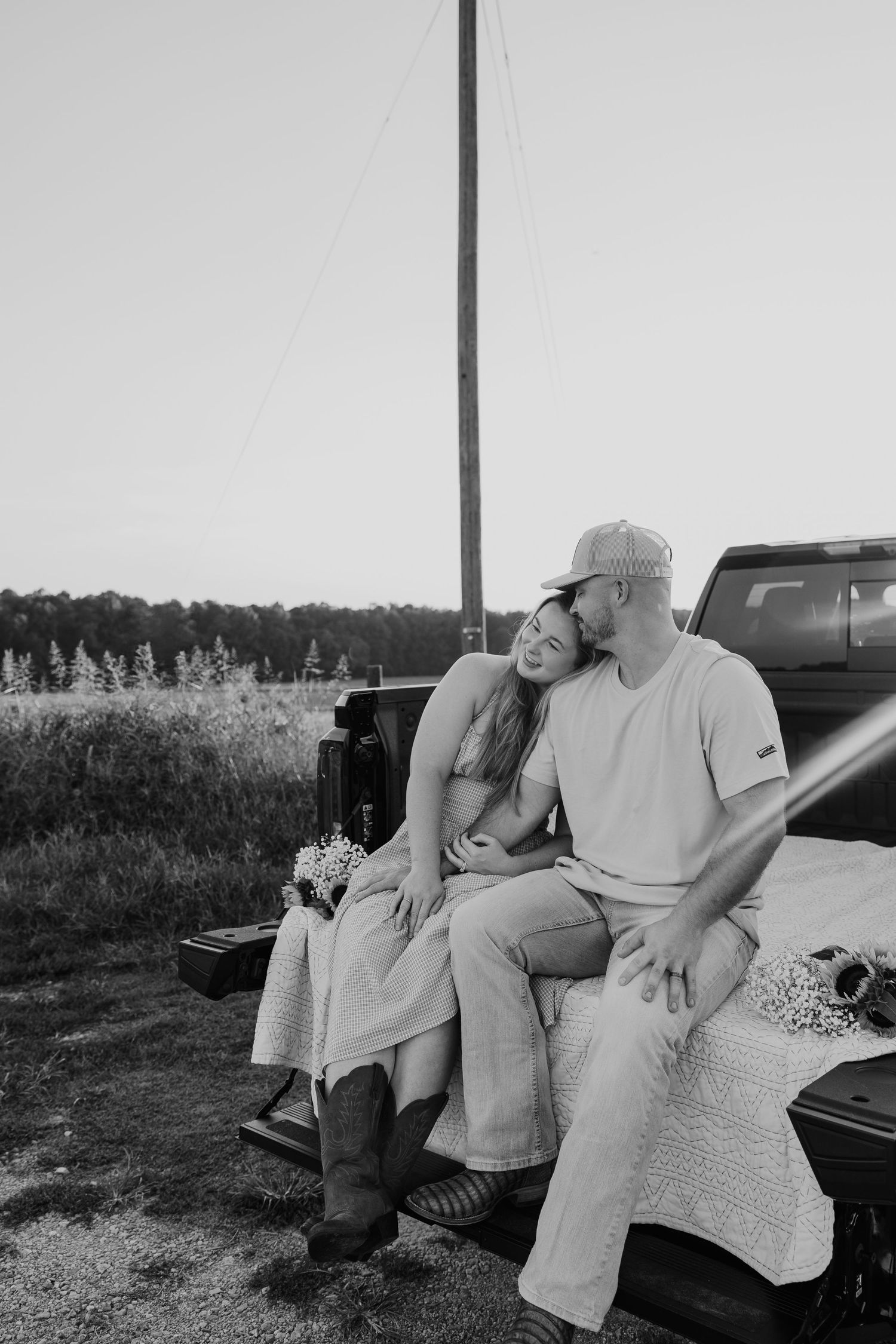 Black and white photo of a couple sitting on a truck tailgate in a field, sharing a tender kiss at dusk.
