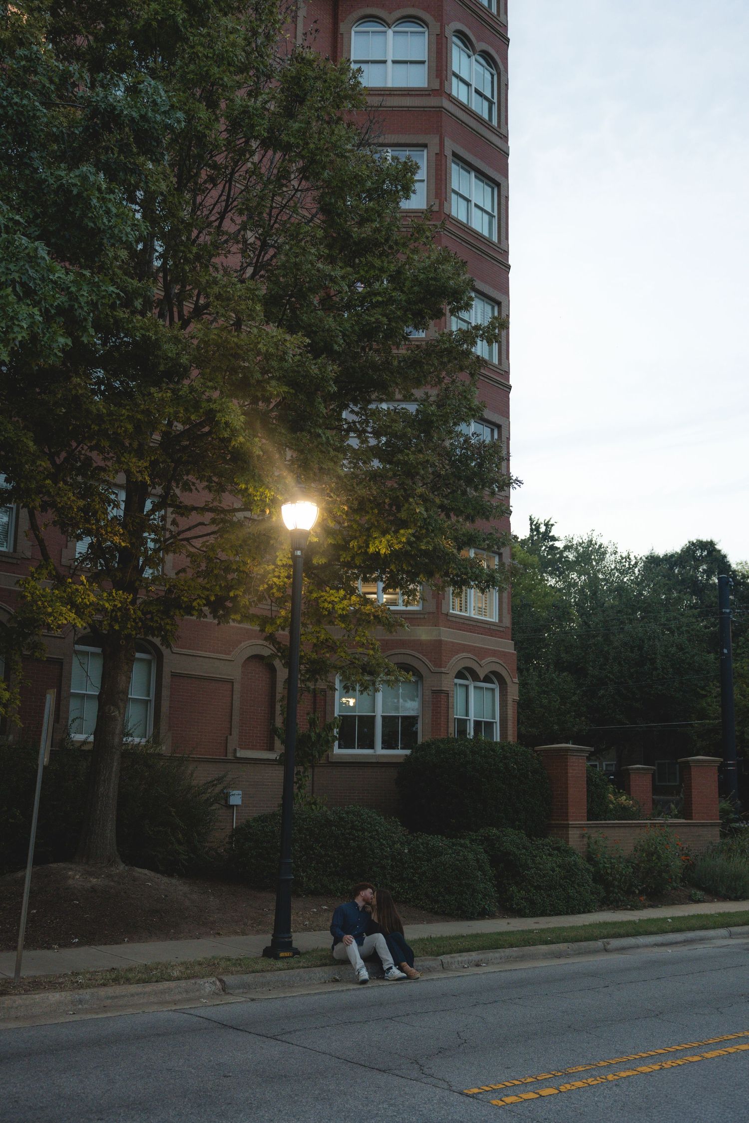 Person sitting under a glowing street lamp beside a tall brick building at dusk on a quiet urban street.