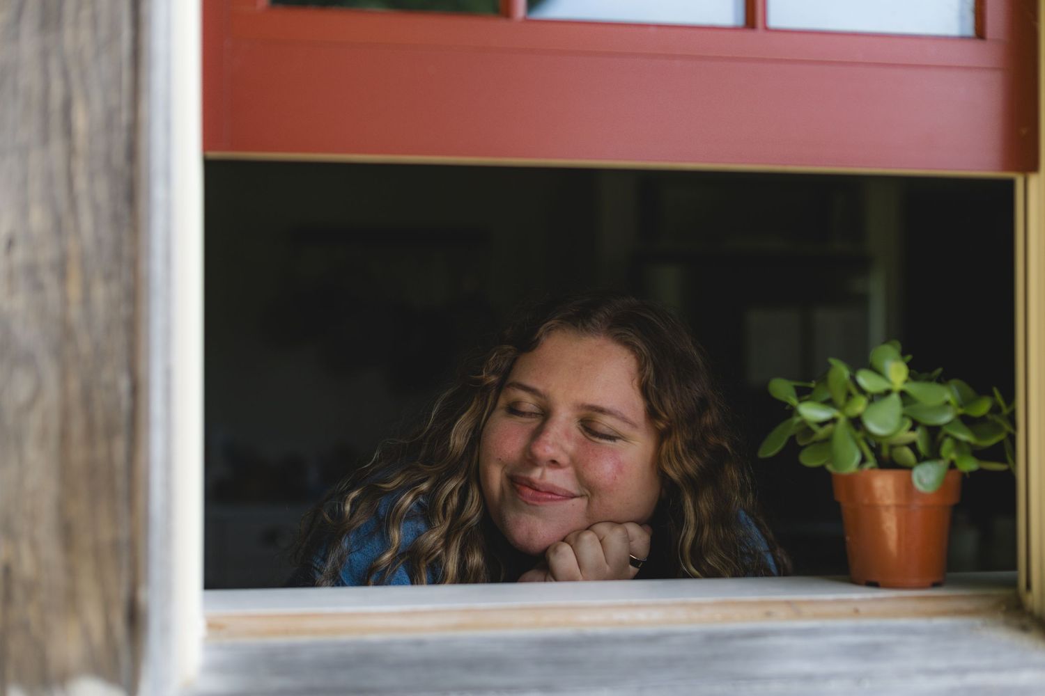 Young girl with curly hair resting her chin on hands, smiling dreamily at a window ledge with a small potted plant.