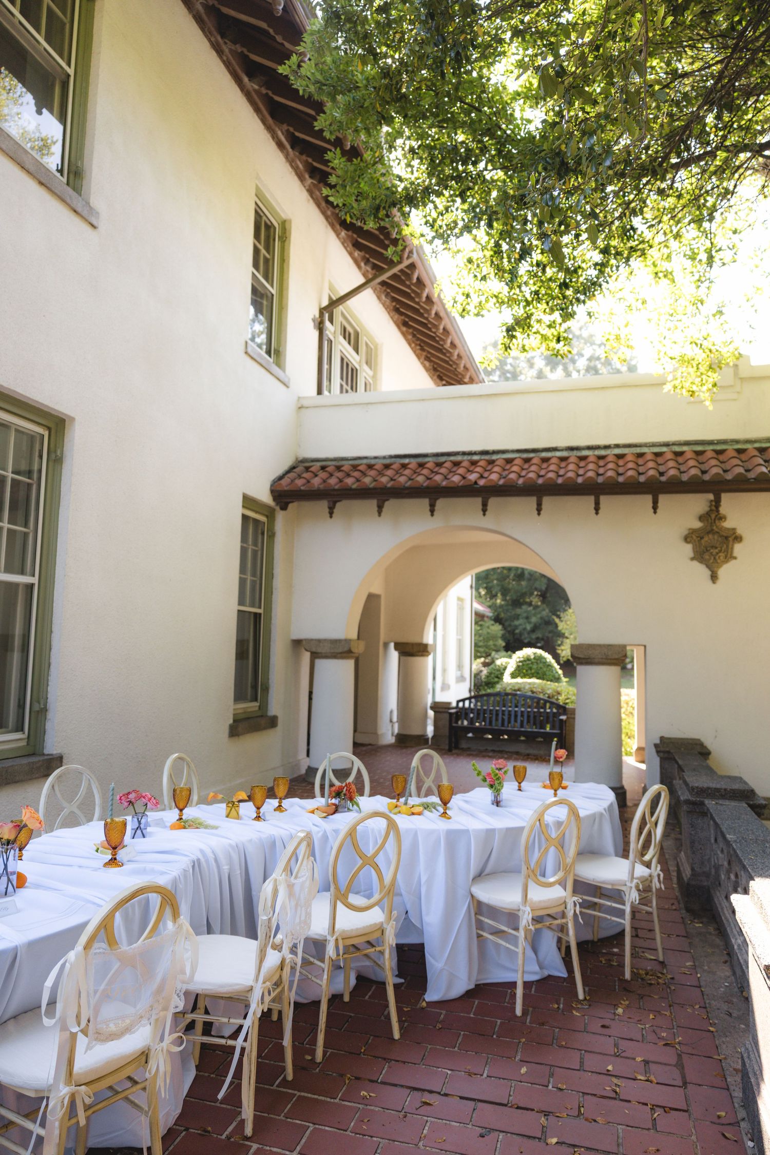 Elegant outdoor dining setup with white tablecloths and gold chairs in a Spanish-style courtyard with arched walkway.