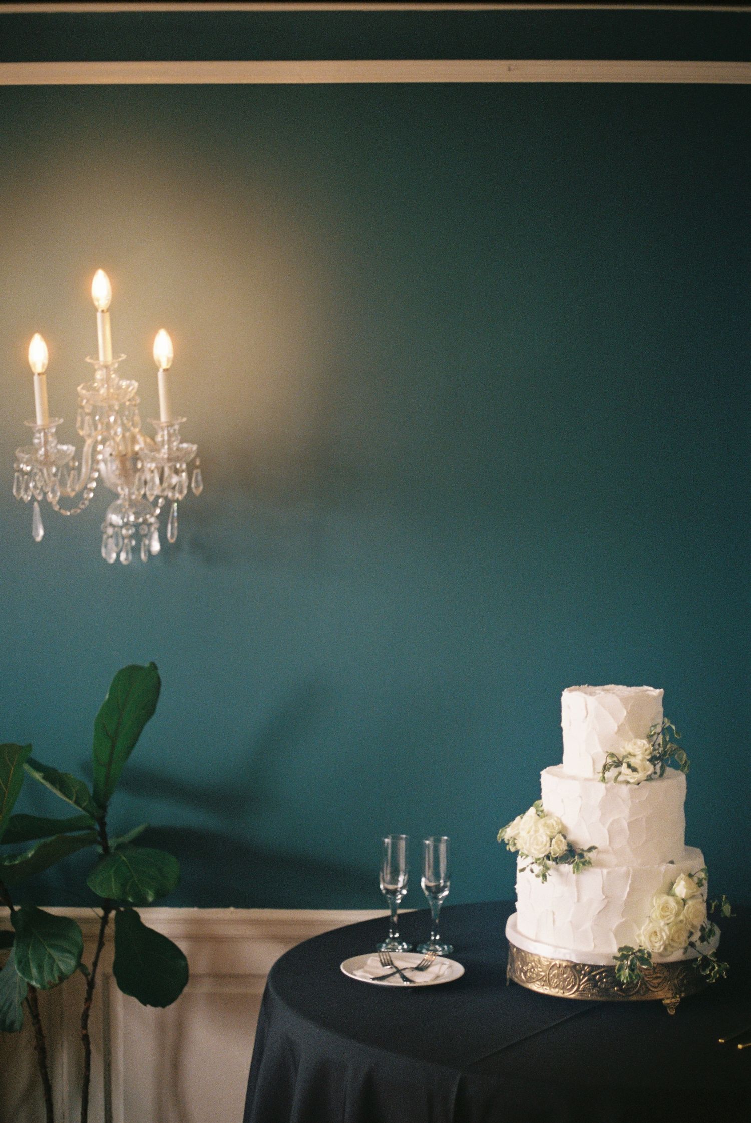 Three-tier white wedding cake with floral accents on black table, crystal wall sconce, and champagne flutes in elegant teal room.