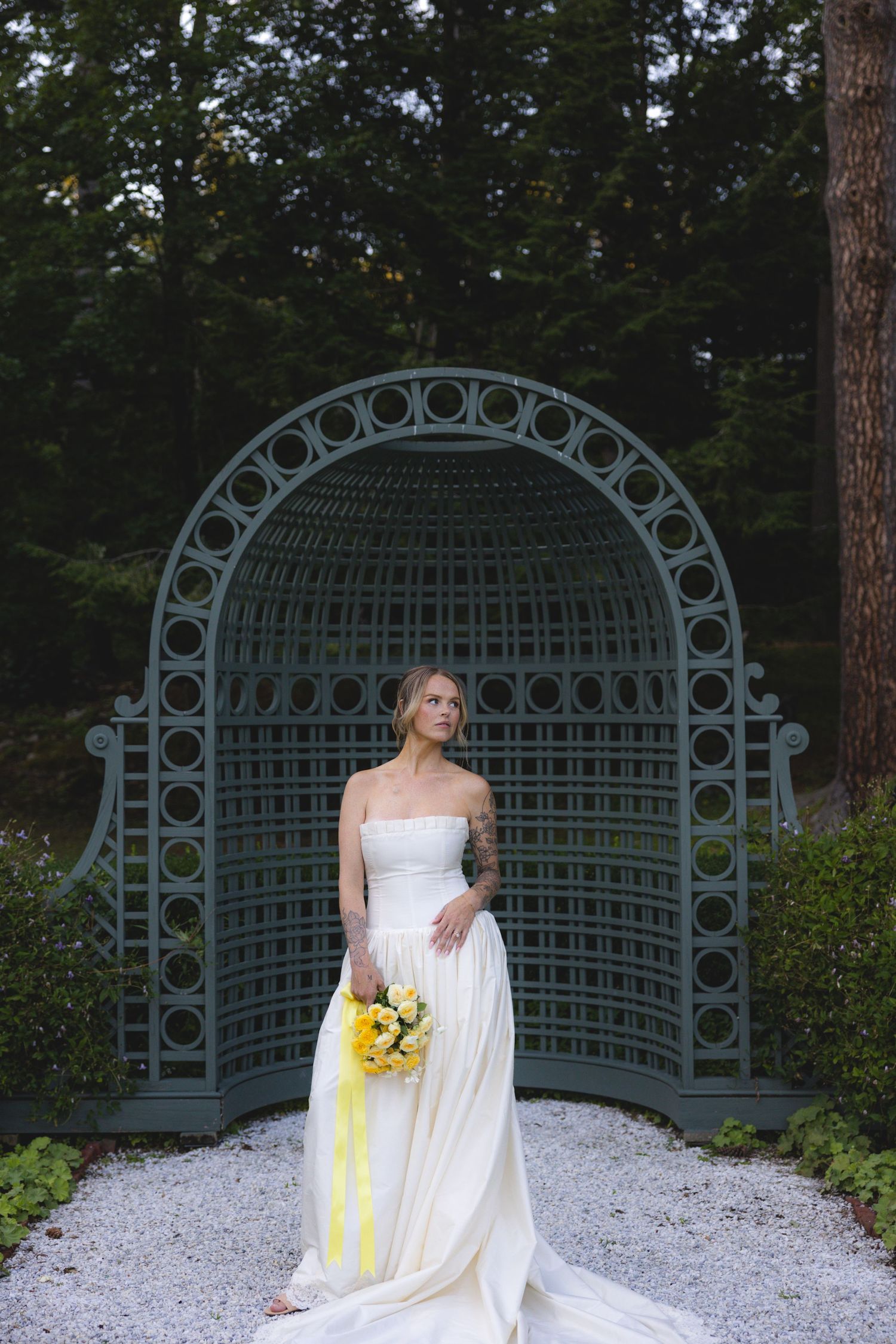 Bride in strapless white gown holding yellow bouquet stands before ornate teal garden arch surrounded by lush greenery.