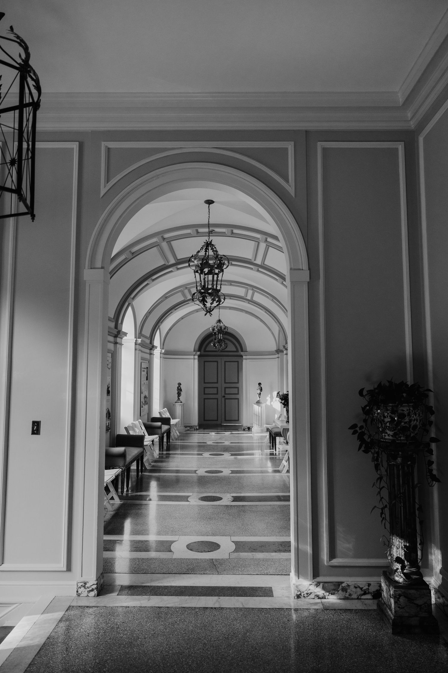 Black and white photo of elegant arched hallway with chandelier lighting and ornate architectural details.