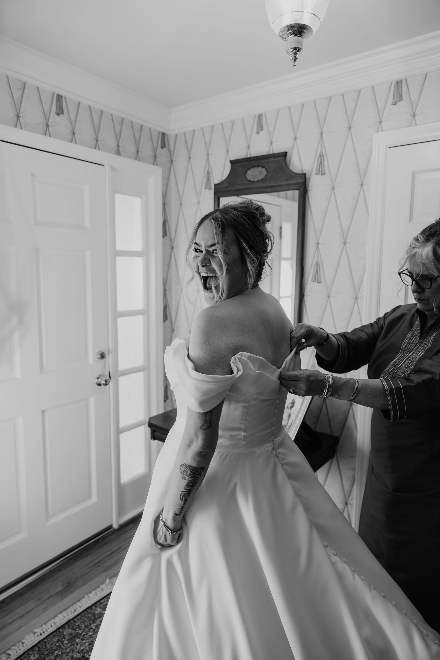 Black and white photo of bride laughing as someone buttons her off-shoulder wedding gown from behind.