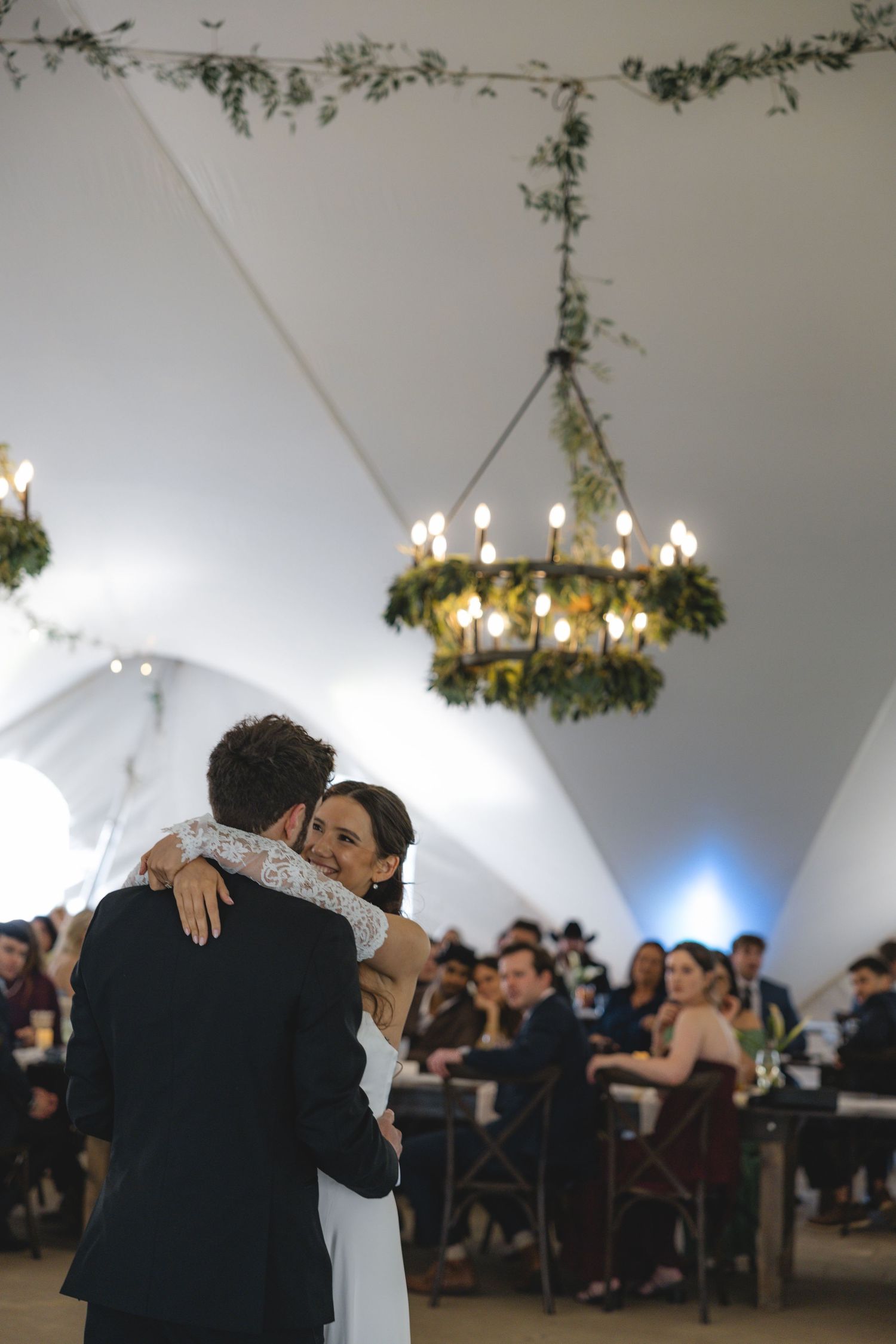 Bride and groom share emotional first dance under floral chandelier in elegant white wedding tent.