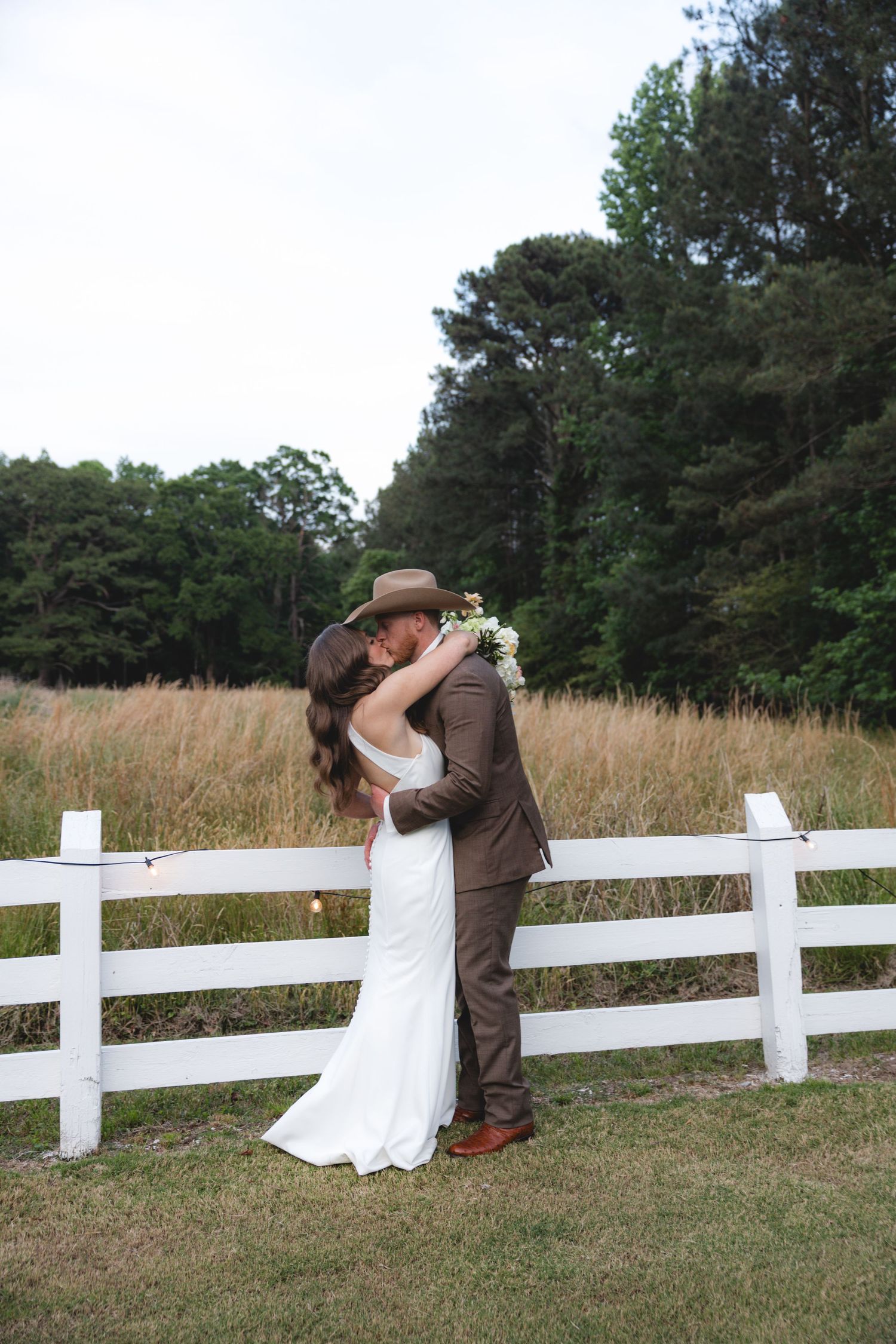 Bride and groom sharing a kiss against a white fence with a golden field and pine trees in the background.