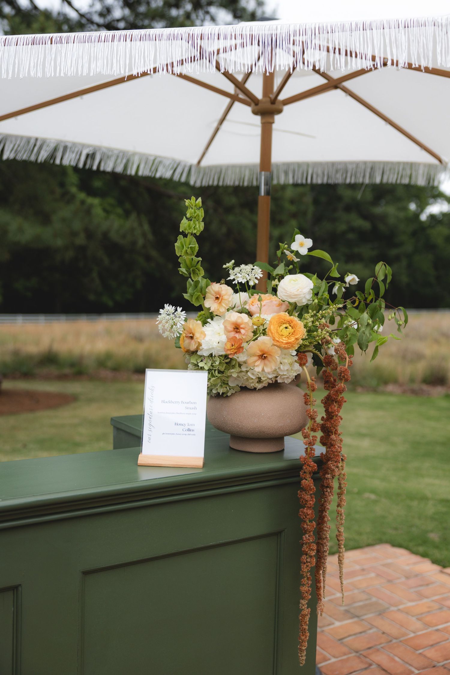 Peach and white floral arrangement in terracotta urn on green bar cart under fringed umbrella.