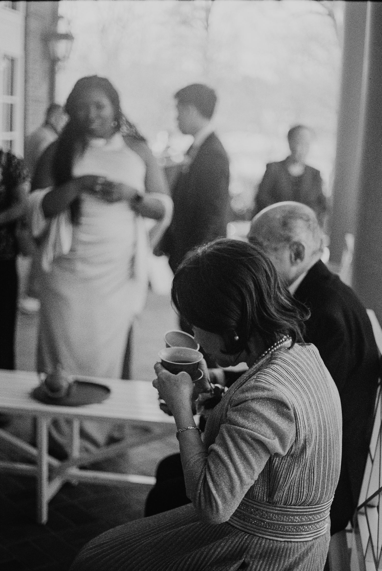 Black and white photo of wedding guests mingling outdoors, woman sipping from teacup in foreground.