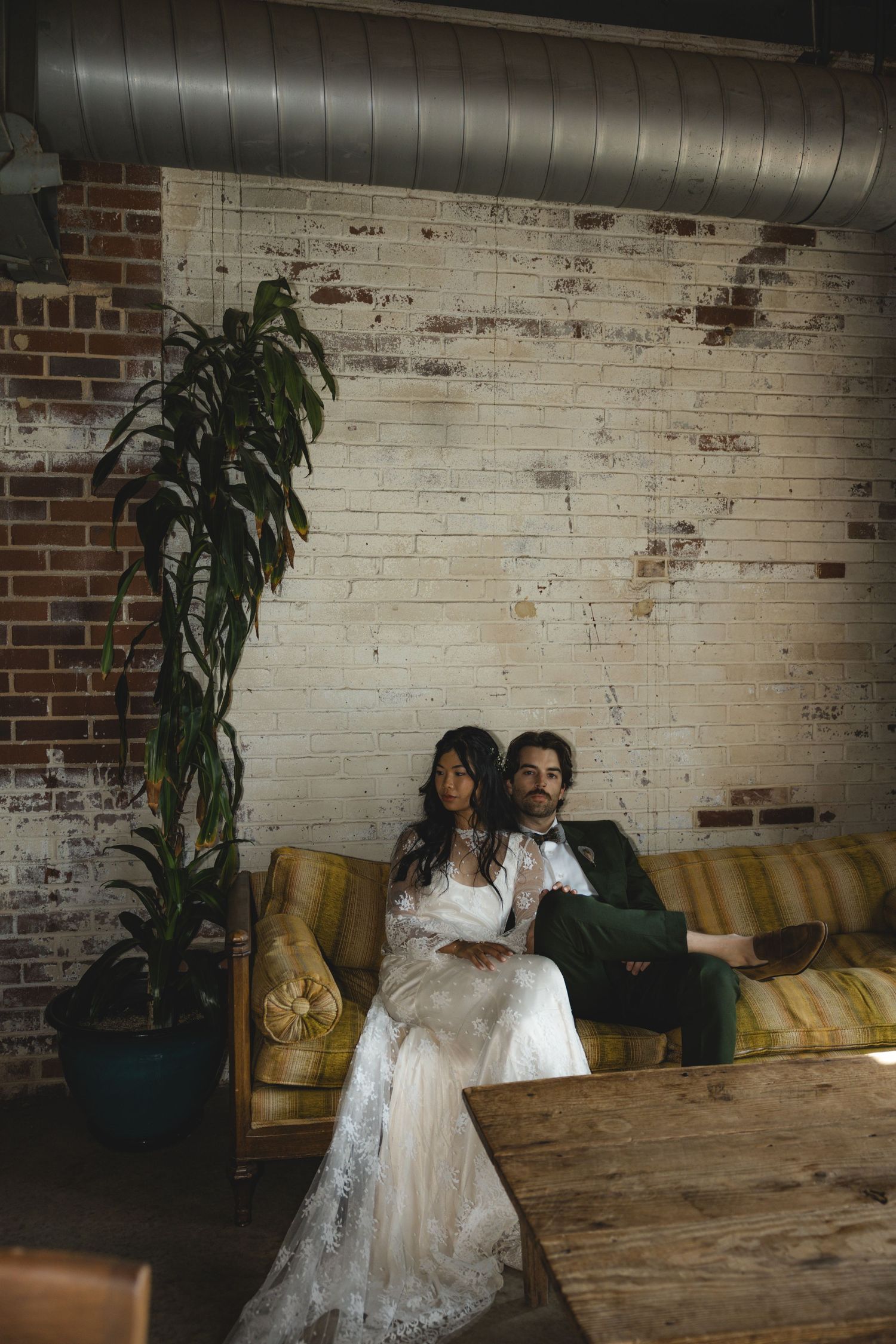Bride and groom sitting together on a vintage yellow sofa against a whitewashed brick wall in an industrial wedding venue.