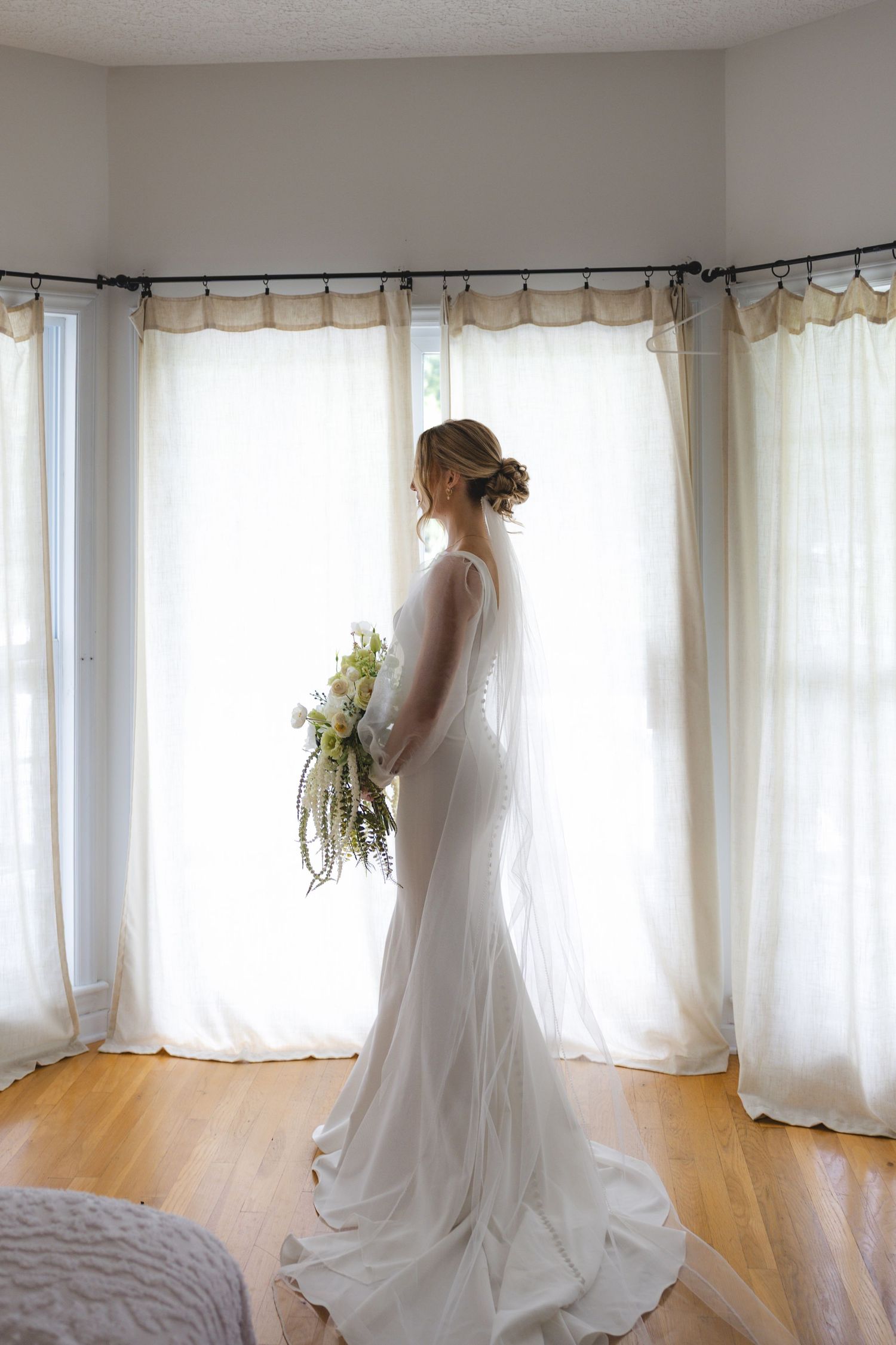 Bride in white wedding gown holding bouquet stands by sunlit windows with sheer curtains on hardwood floor.