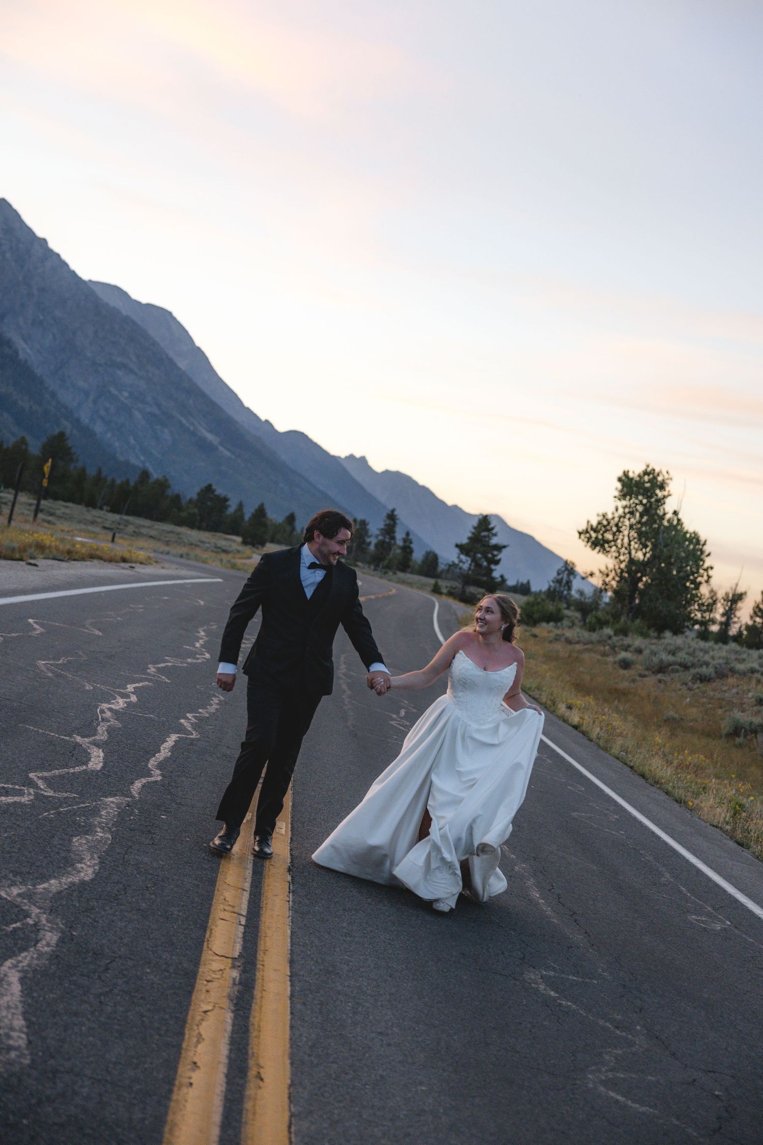 Bride and groom walk hand-in-hand down a mountain road at sunset in Grand Teton National Park.