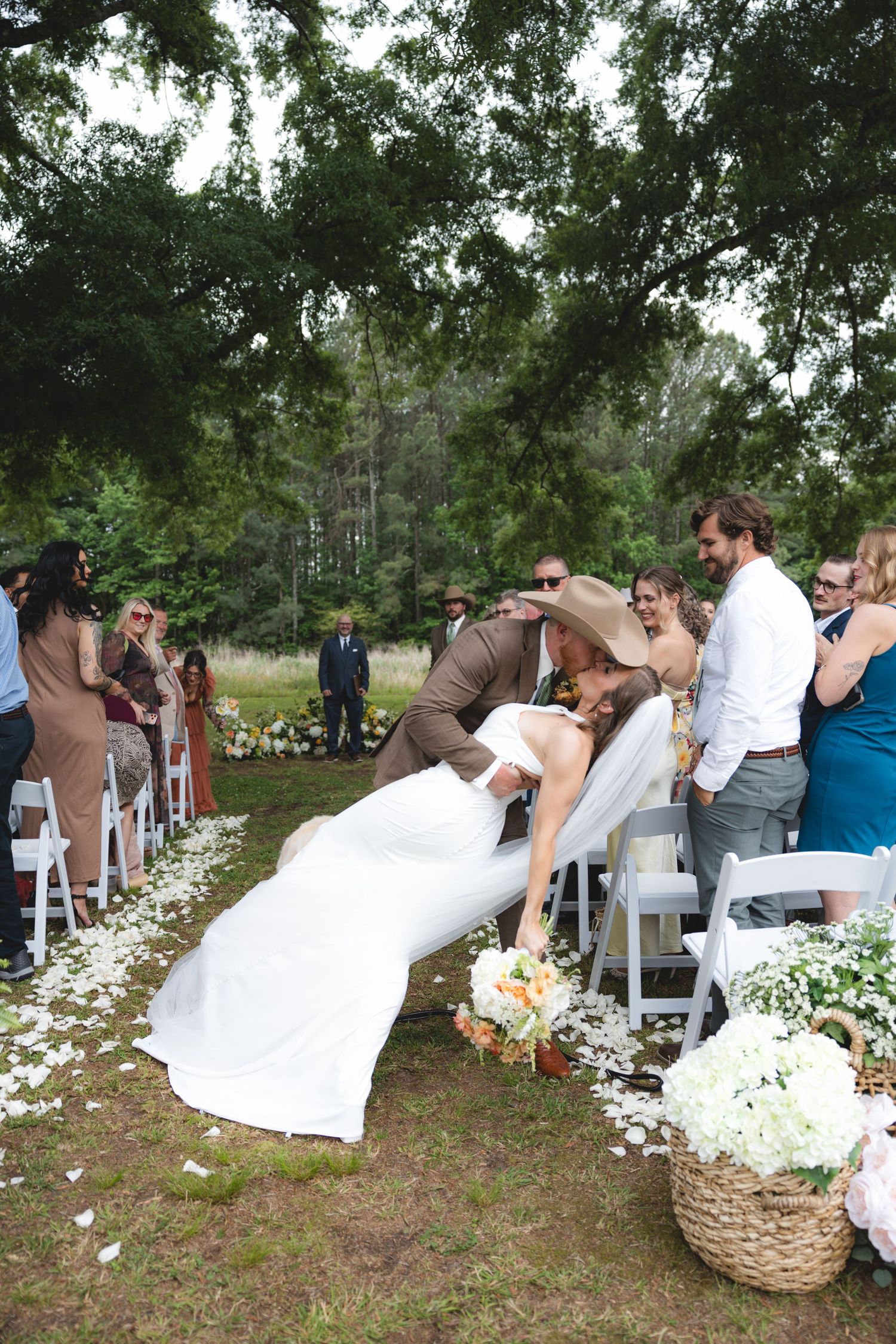 Groom in cowboy hat dips bride in white gown during outdoor wedding ceremony aisle lined with white petals.