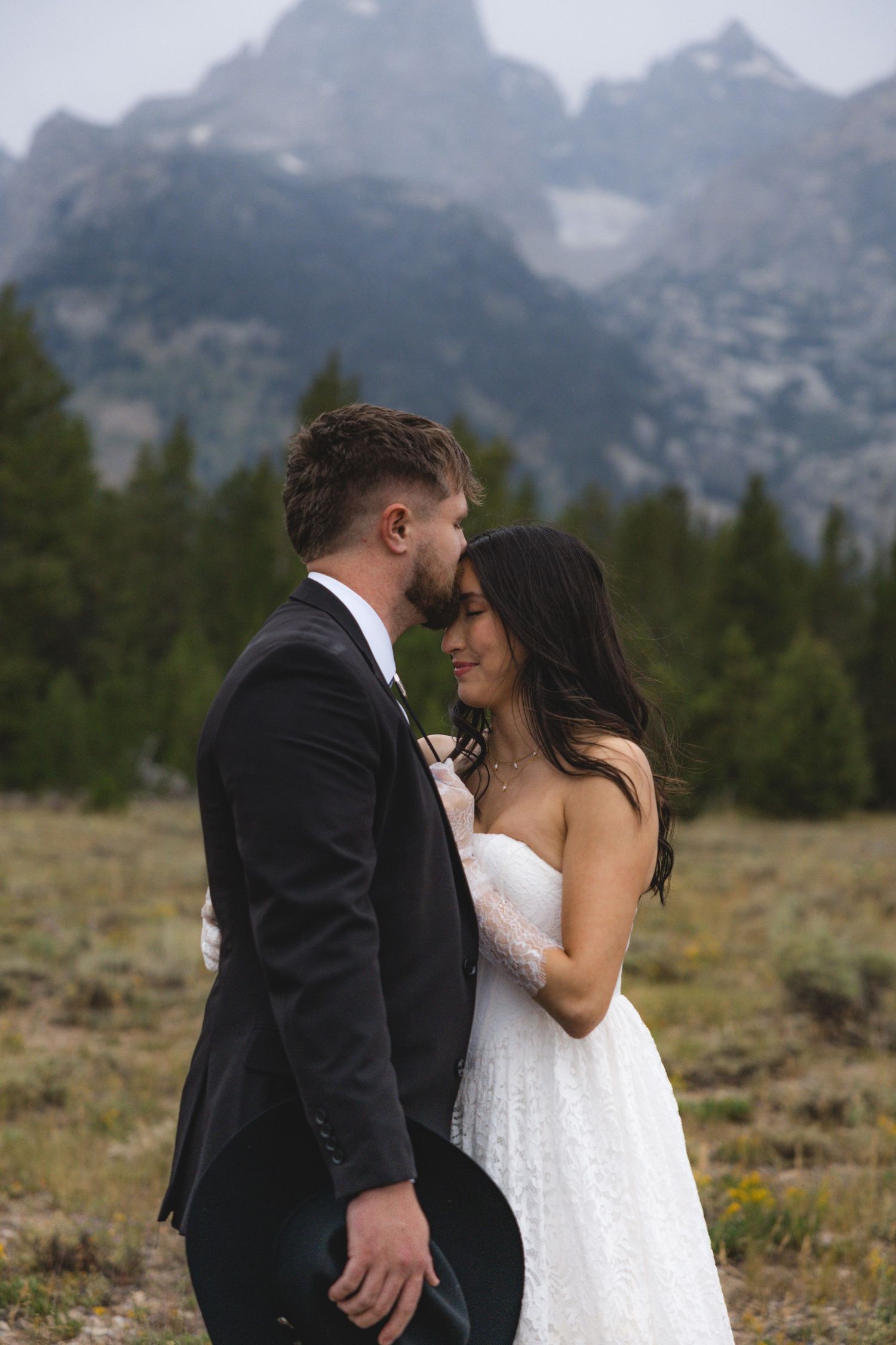 A couple shares a romantic moment in wedding attire with the Grand Teton mountains and pine trees in the background.