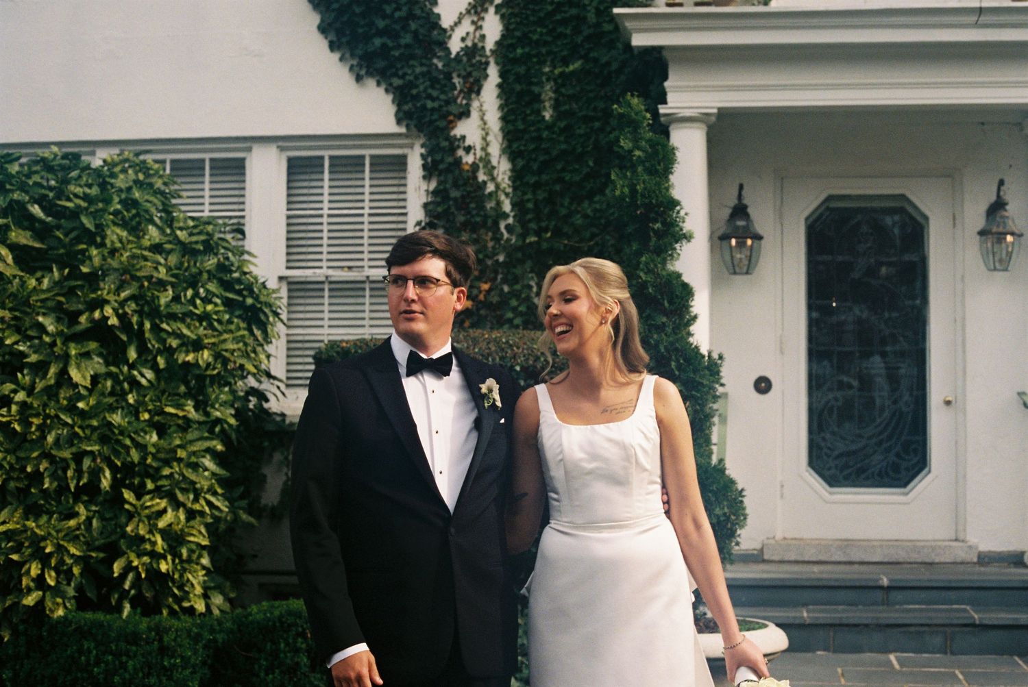 A bride and groom stand together in front of a white building with greenery surrounding them.