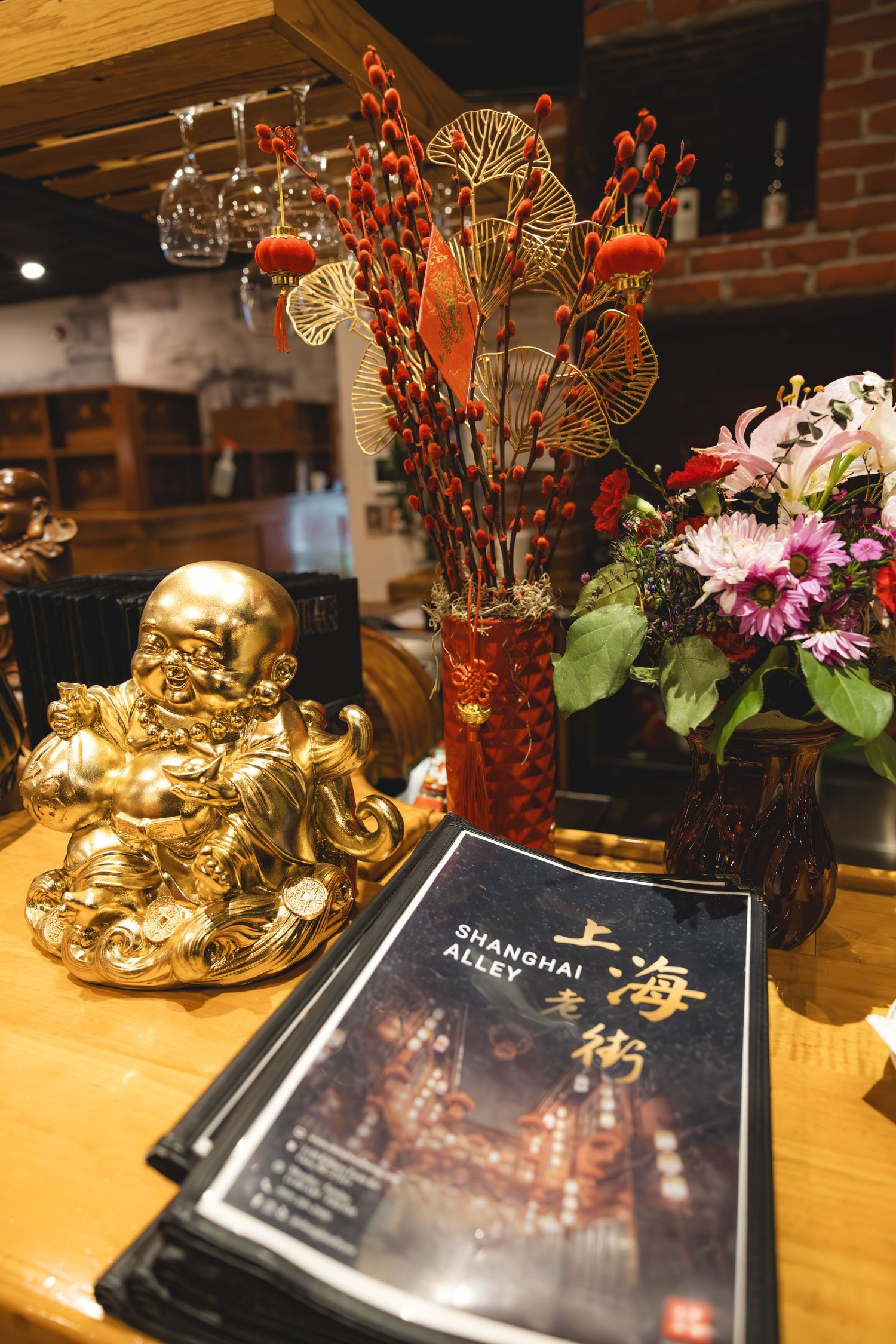 Golden Buddha statue beside Shanghai Alley restaurant menus, red vase with decorative branches, and fresh flowers on wooden counter.