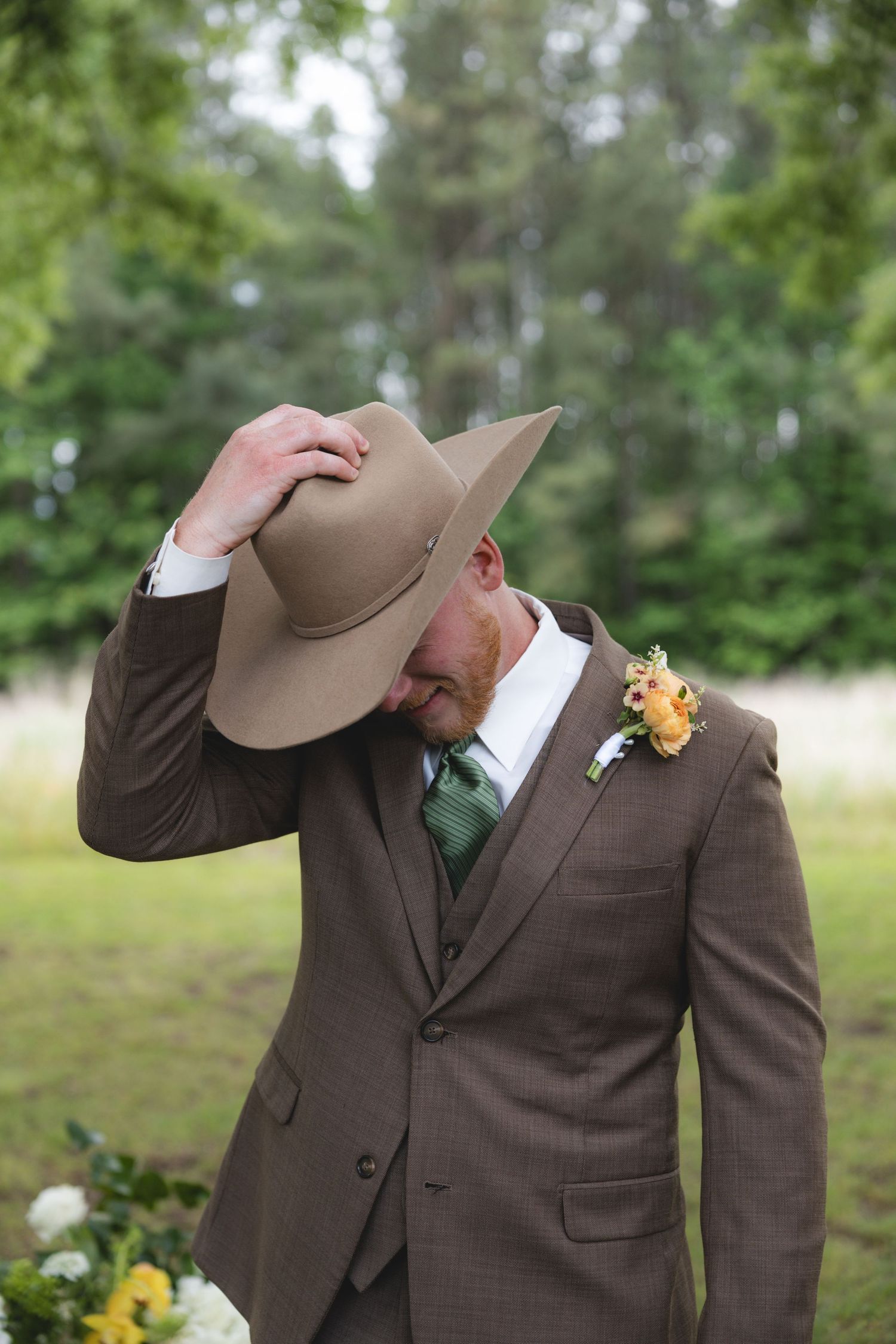 Groom in brown three-piece suit with green tie and floral boutonniere tips tan cowboy hat in a field.