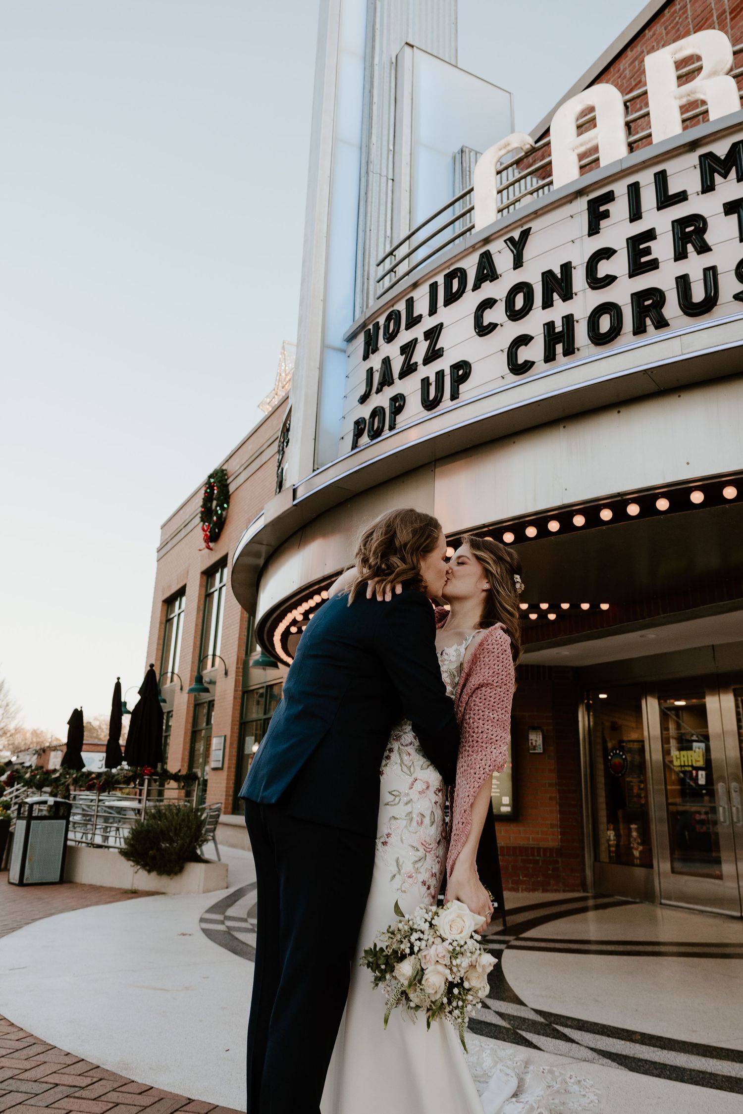 Bride and groom share a kiss outside a classic theater marquee displaying holiday films and jazz concerts.