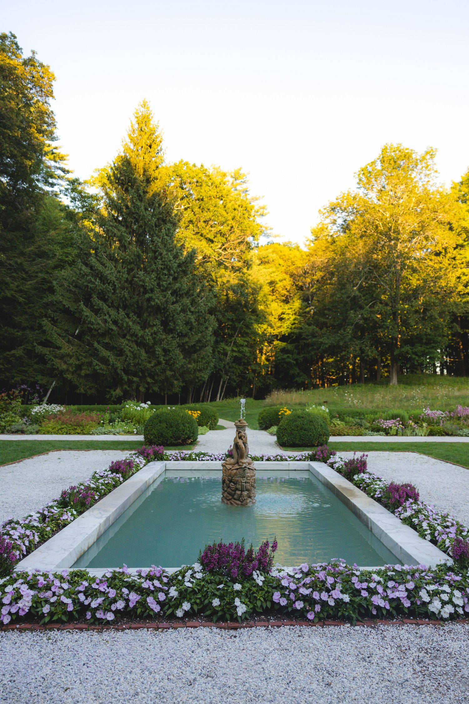 Formal garden fountain surrounded by colorful flowers and lush green trees in a manicured landscape at dusk.
