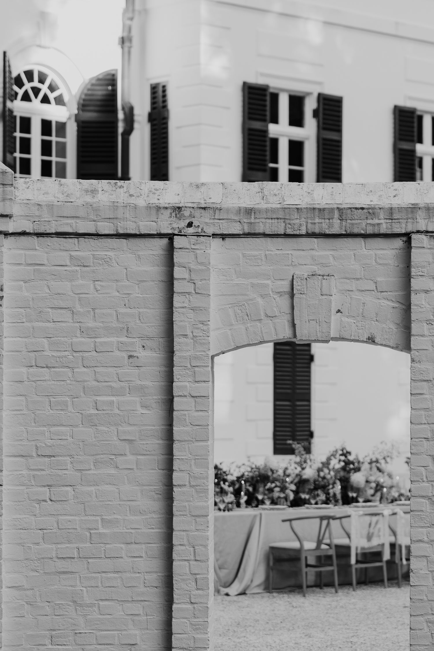 Black and white photo of a brick archway revealing an elegant outdoor dining setup with floral arrangements and chairs.