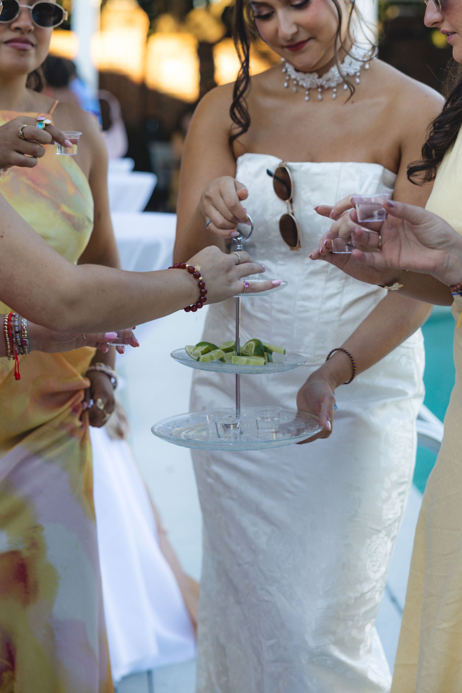 Bride in white strapless gown serving appetizers on tiered tray at outdoor wedding reception with guests.