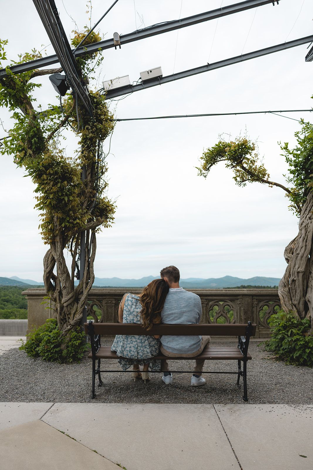 Couple sitting on a bench under a pergola with ivy-covered trees, overlooking a scenic mountain landscape.