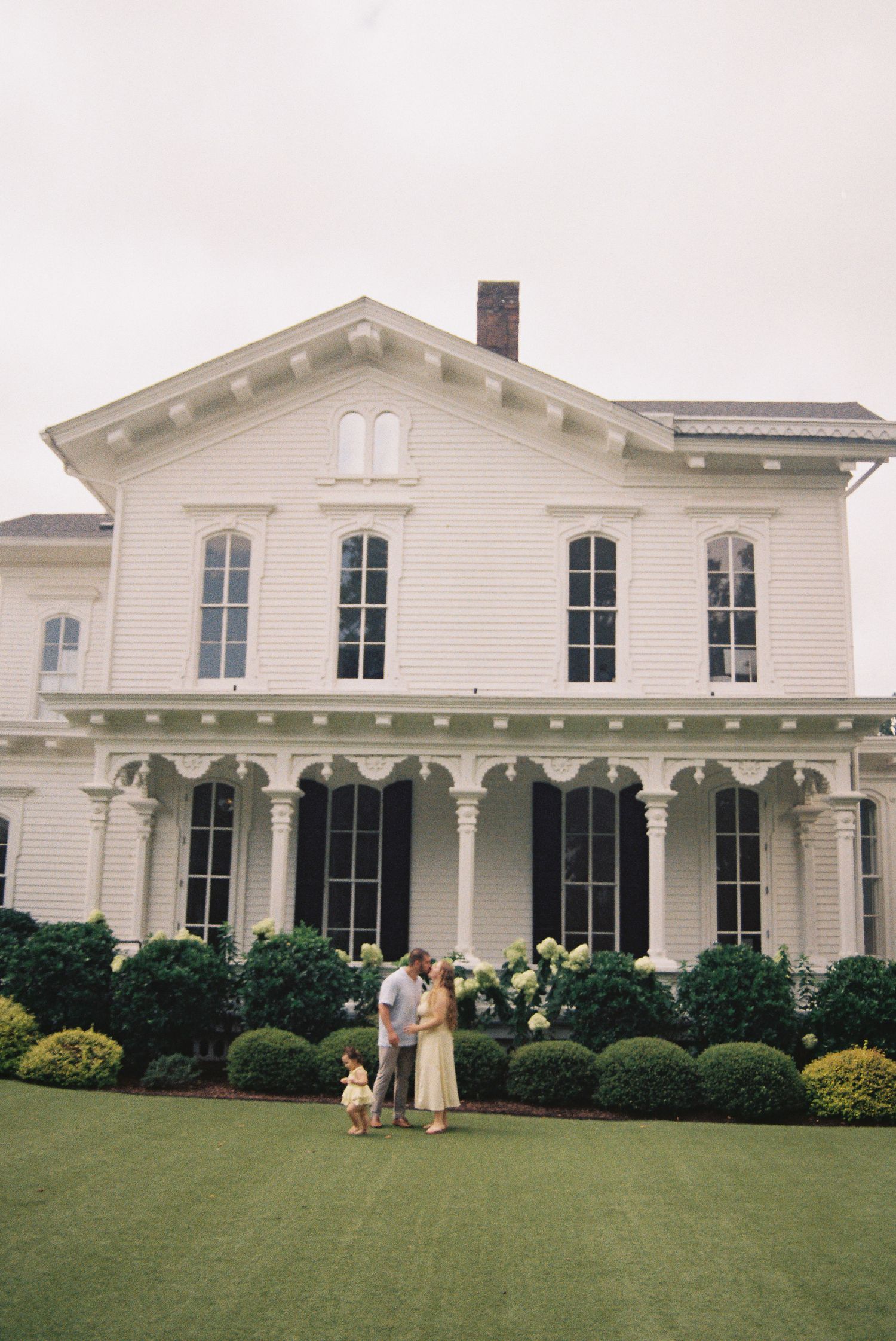 Family posing on lawn of elegant white Southern mansion with columns, manicured hedges, and hydrangeas.