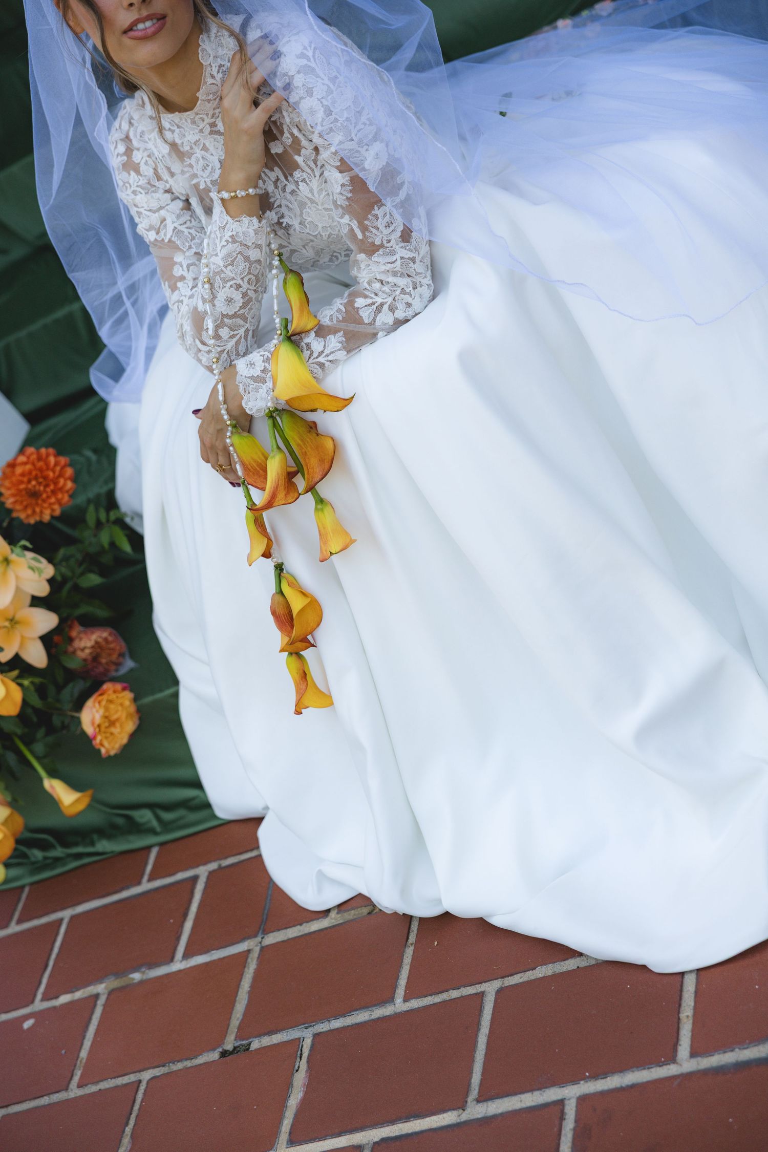 Bride in white lace gown holding cascading yellow calla lily bouquet on brick steps with orange floral arrangements.