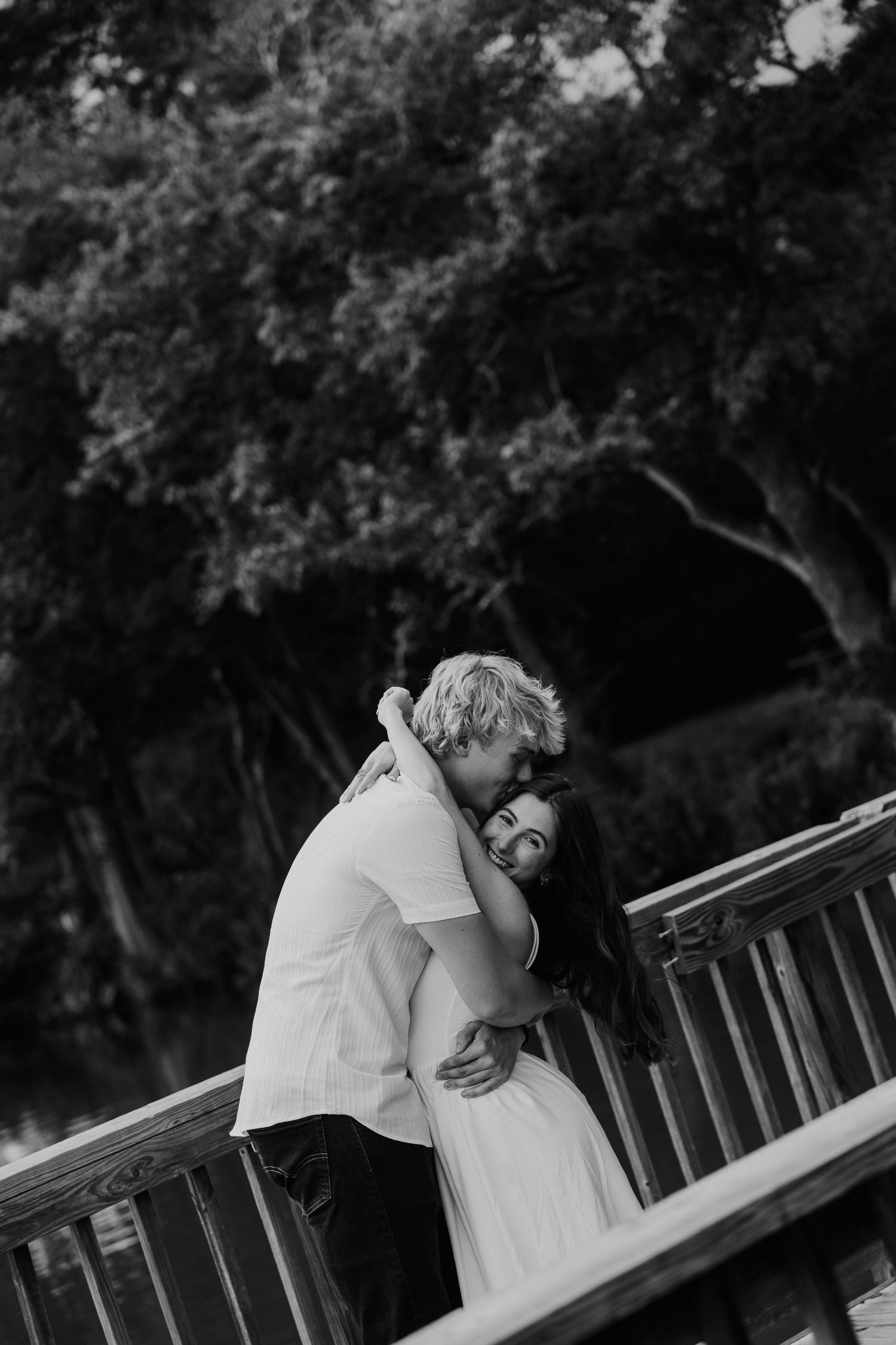 Black and white photo of a couple embracing tenderly on a wooden bridge surrounded by lush trees.