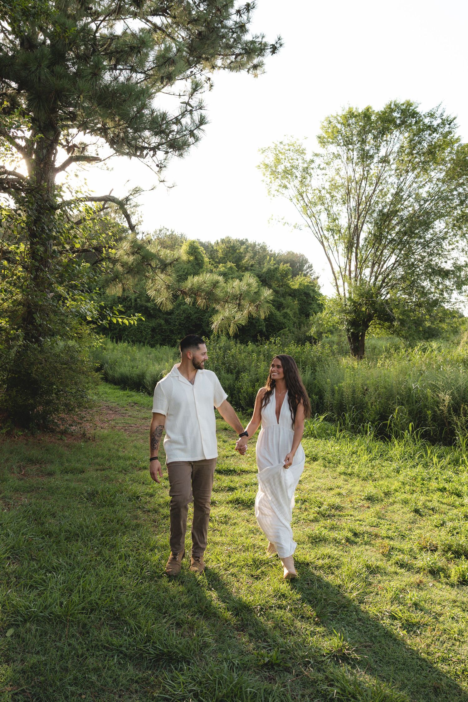 Couple in white outfits holding hands while walking on a lush green path surrounded by trees.